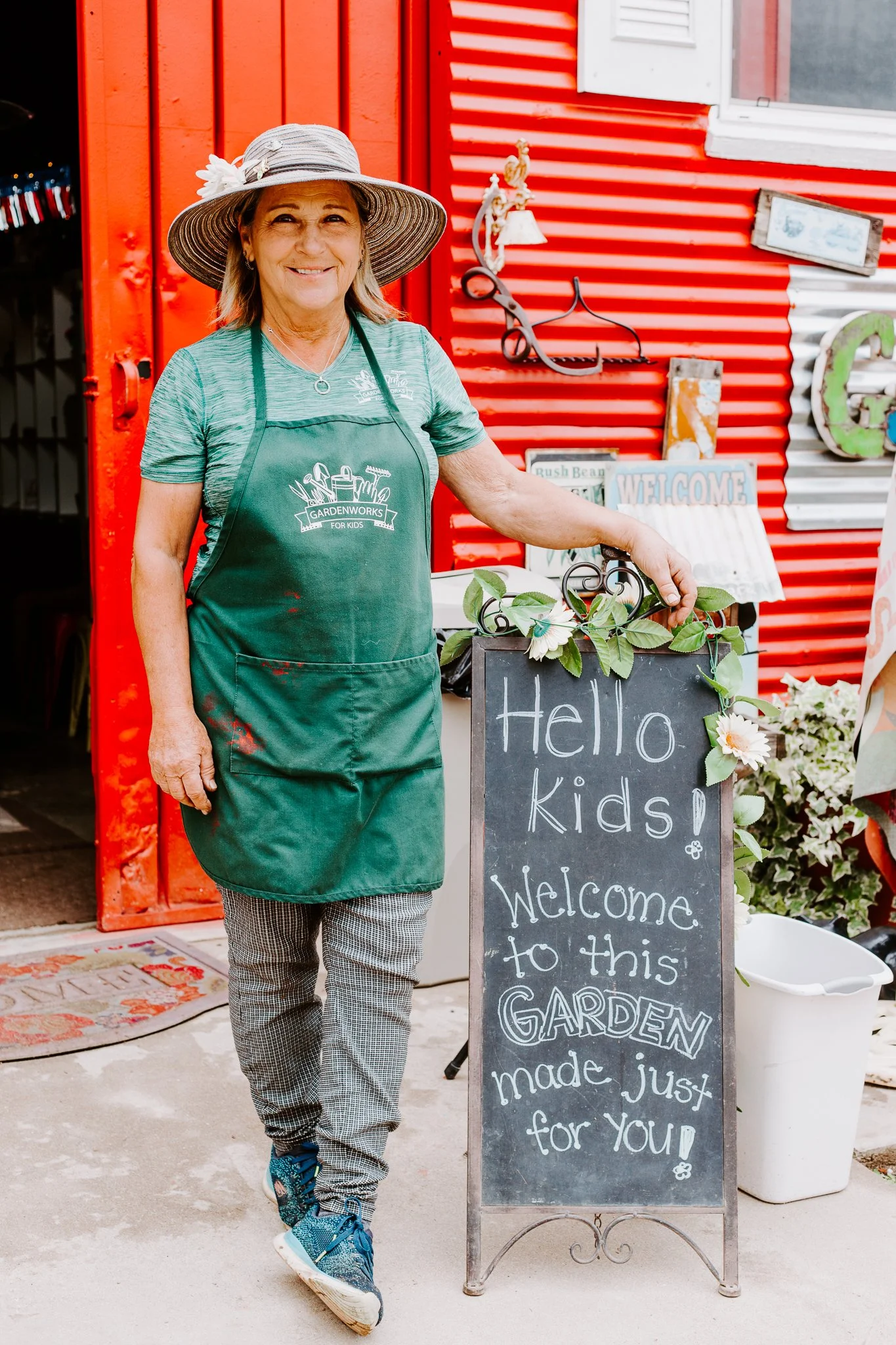 A smiling woman in a green apron and a large sun hat stands outside a red building, holding a small blackboard with a message about a garden made just for kids.