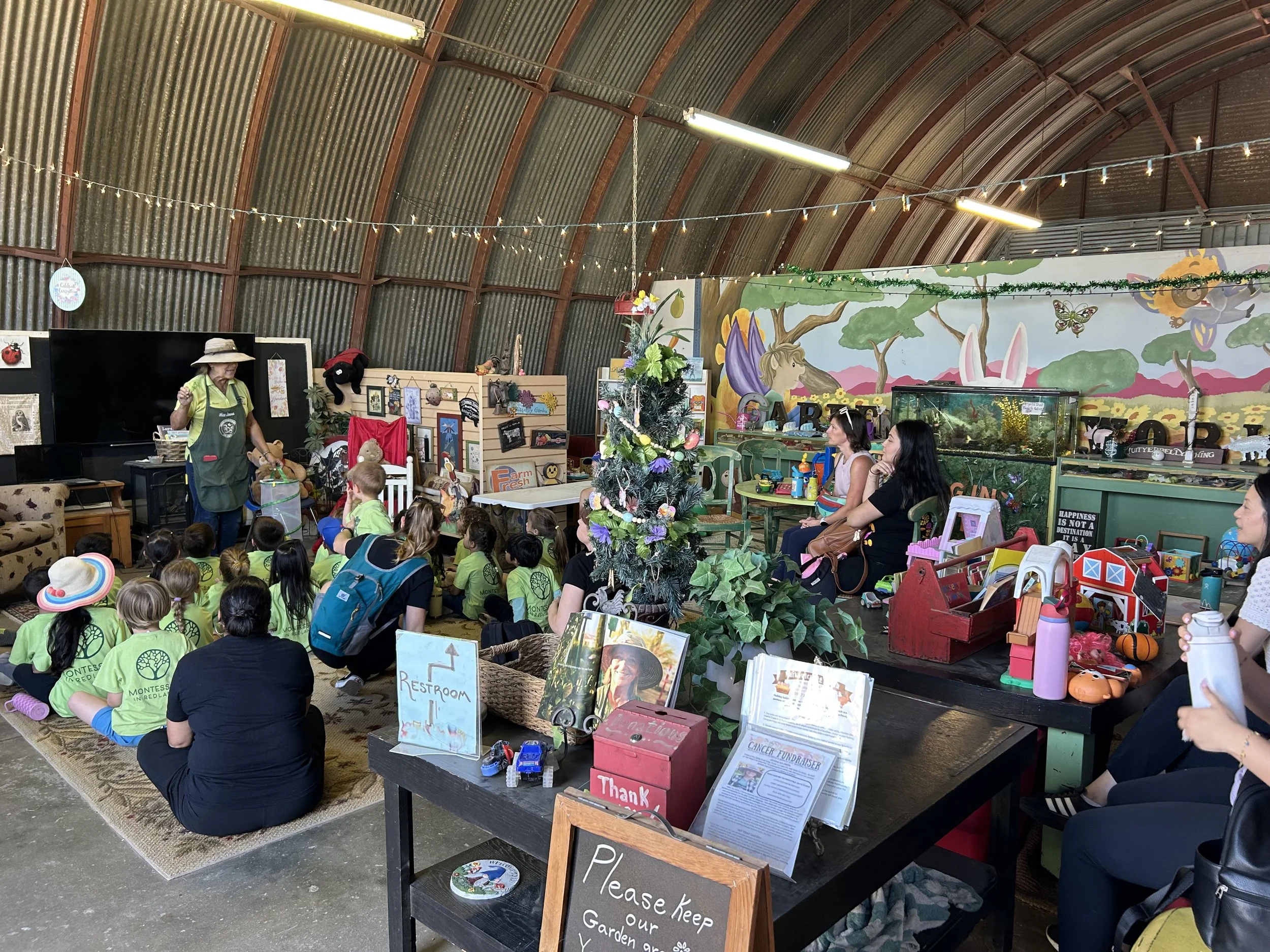 A gathering of children and adults in a decorated indoor space with a colorful mural, string lights, and various tables displaying items and signs, including a Christmas tree in the center.