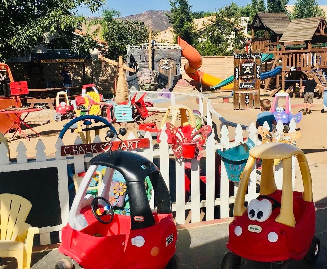 Colorful children's play area with slides, small cars, toys, and a wooden fence, surrounded by trees and a clear blue sky.
