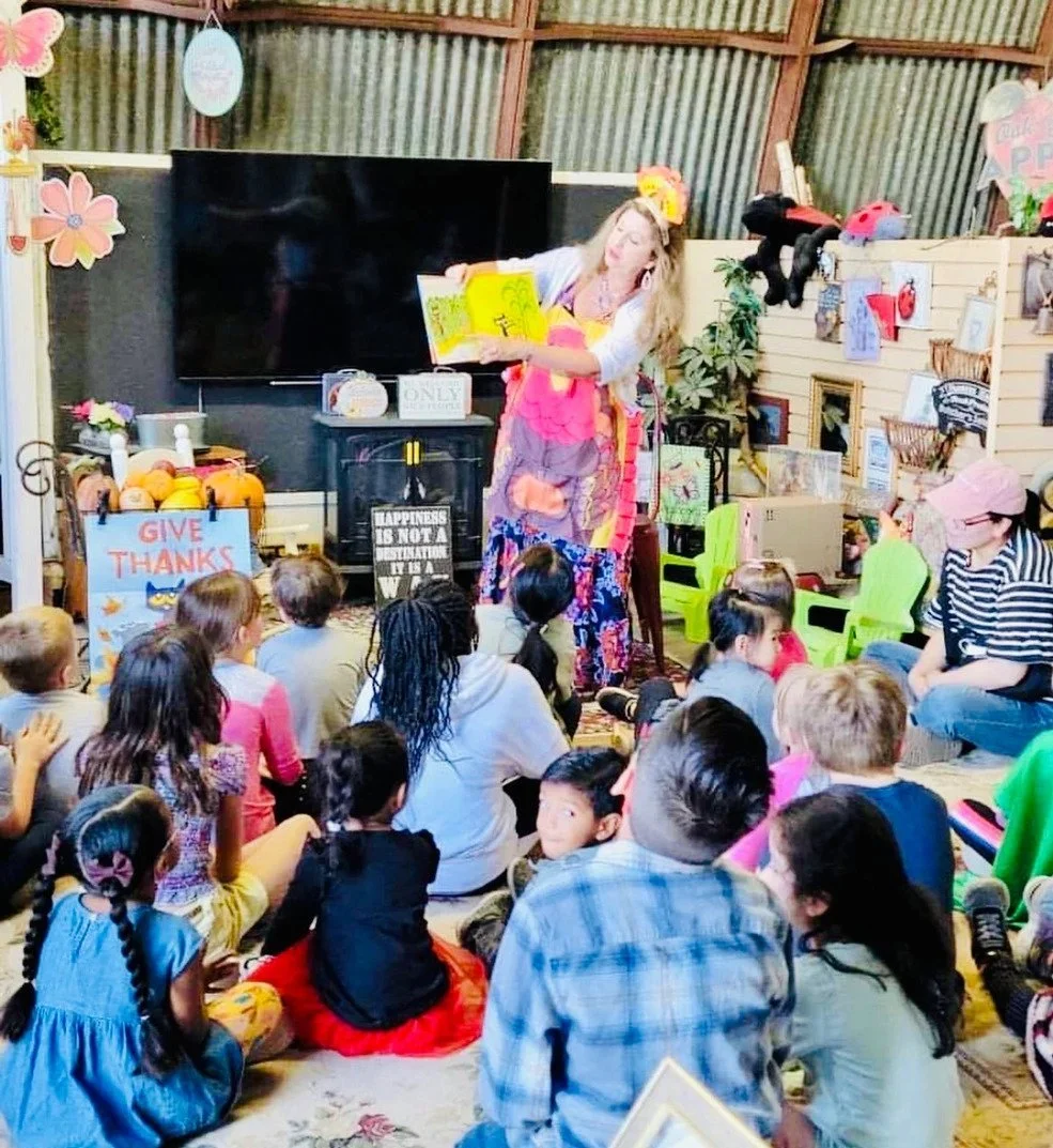 A woman dressed as a clown or performer reads a colorful book to a group of children gathered on the floor in a cozy, decorated indoor space.