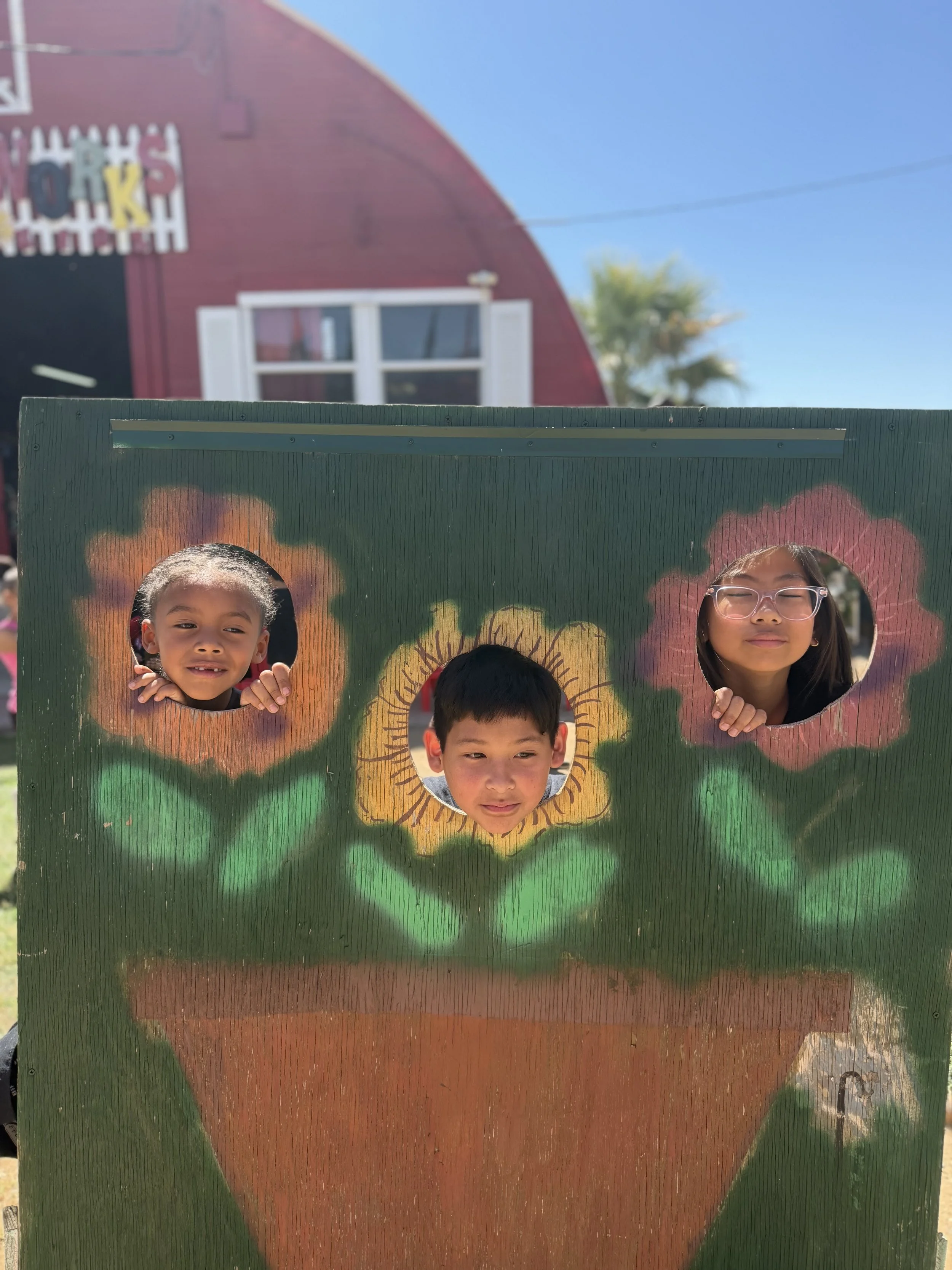 Three children peeking through heart-shaped cutouts in a colorful painted wooden board outdoors, with a red building and blue sky in the background.