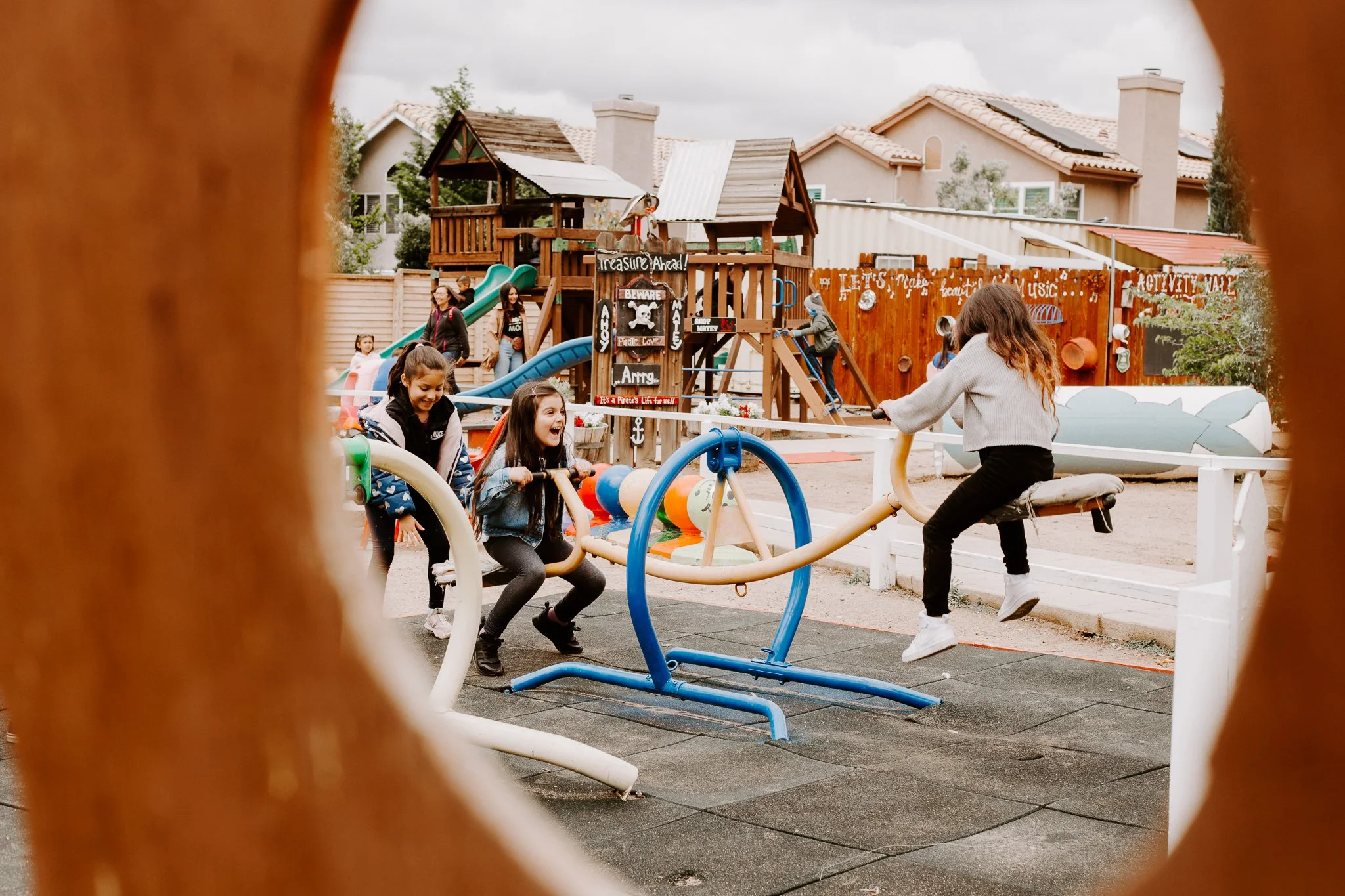 Children playing on playground equipment including a slide and a see-saw at an outdoor play area, viewed through a gap in a wooden fence.