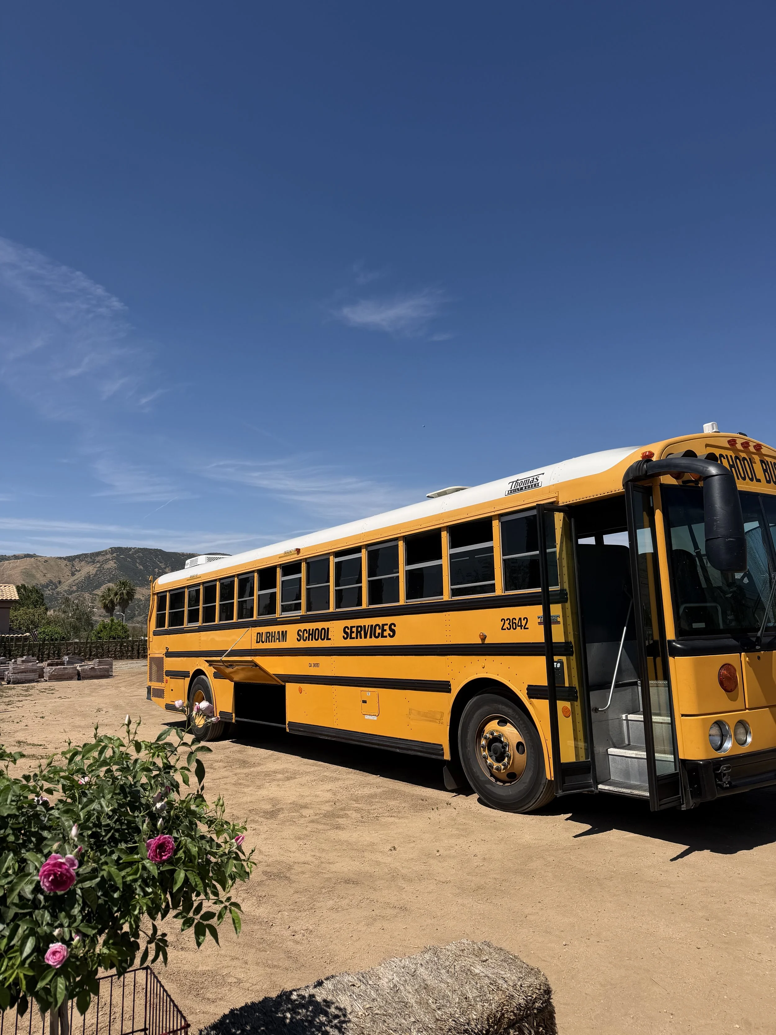 A yellow school bus labeled Durham School Services parked on a dirt area with mountains in the background and blue sky overhead. There are pink roses and a tree in the foreground.
