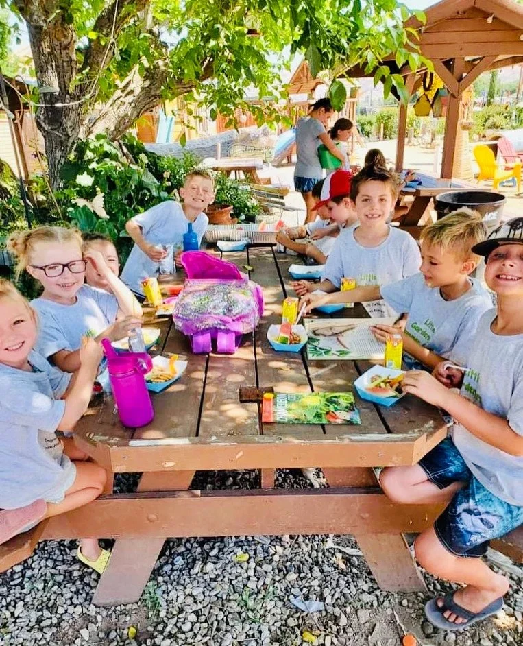 Children enjoying a picnic at an outdoor wooden table under a shaded tree, with some reading and eating, and others standing in the background at a playground.