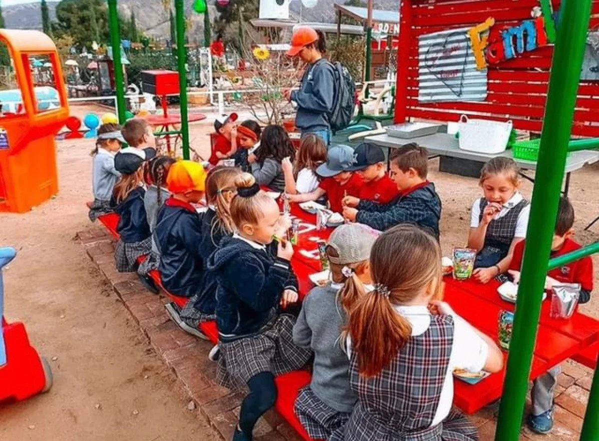 Children dressed in school uniforms sitting at a long red table outdoors, eating together during a school event.