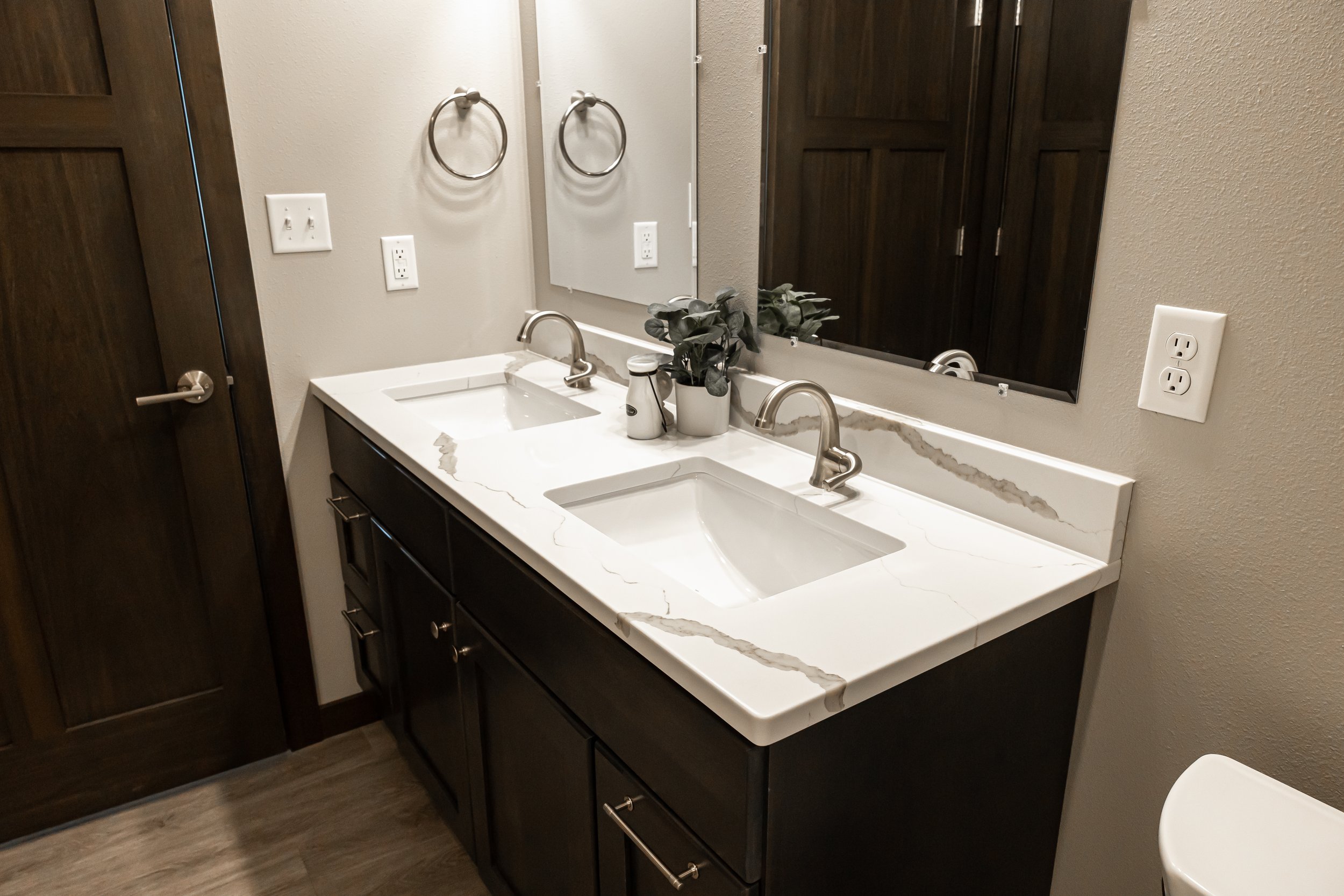 Modern bathroom with double vanity, white countertops, undermount sinks, chrome faucets, wall mirrors, and dark wooden cabinets. Potted plant and toiletries on counter.