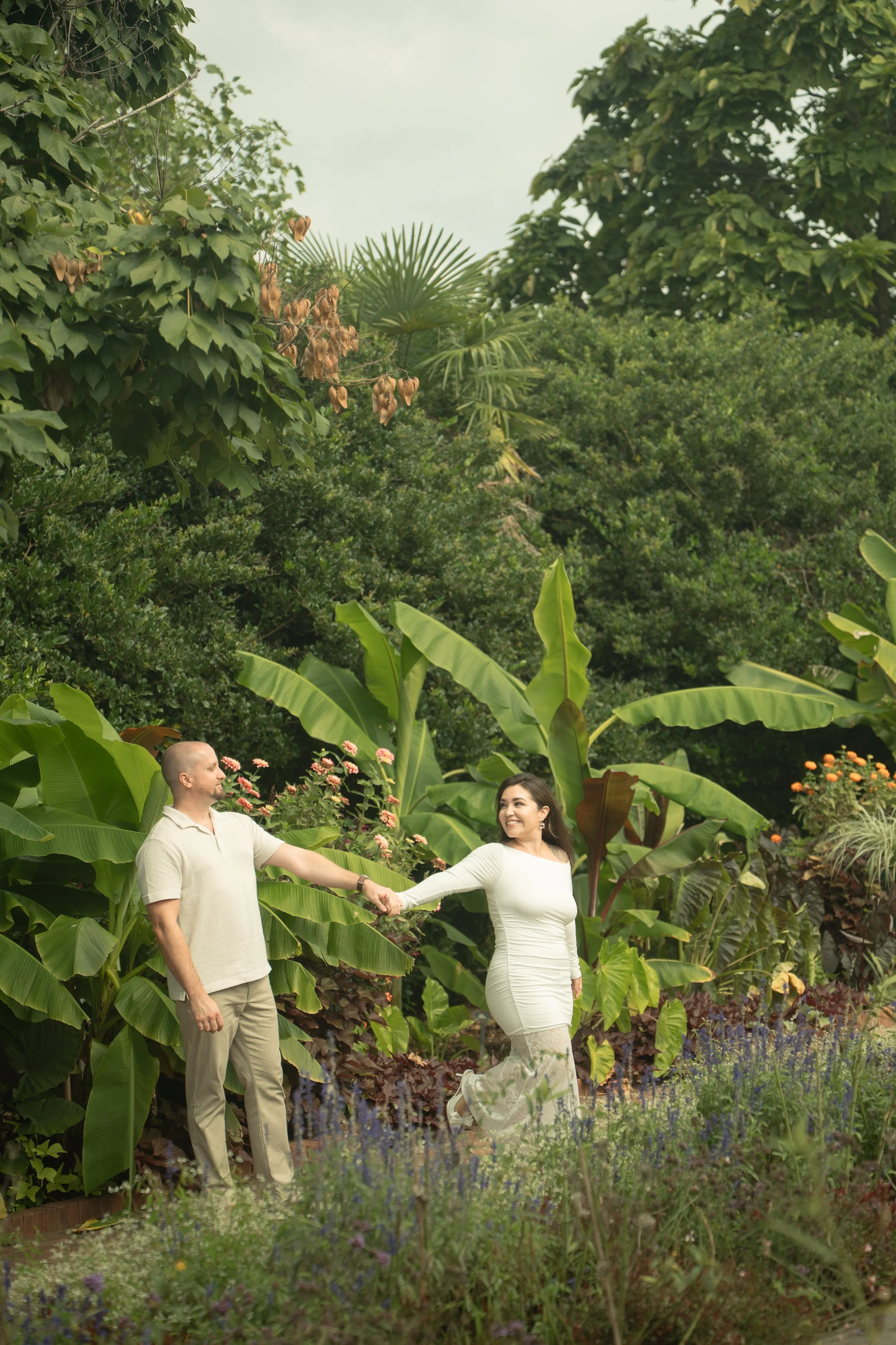 A couple holding hands in a lush garden with large green leaves and vibrant flowers.