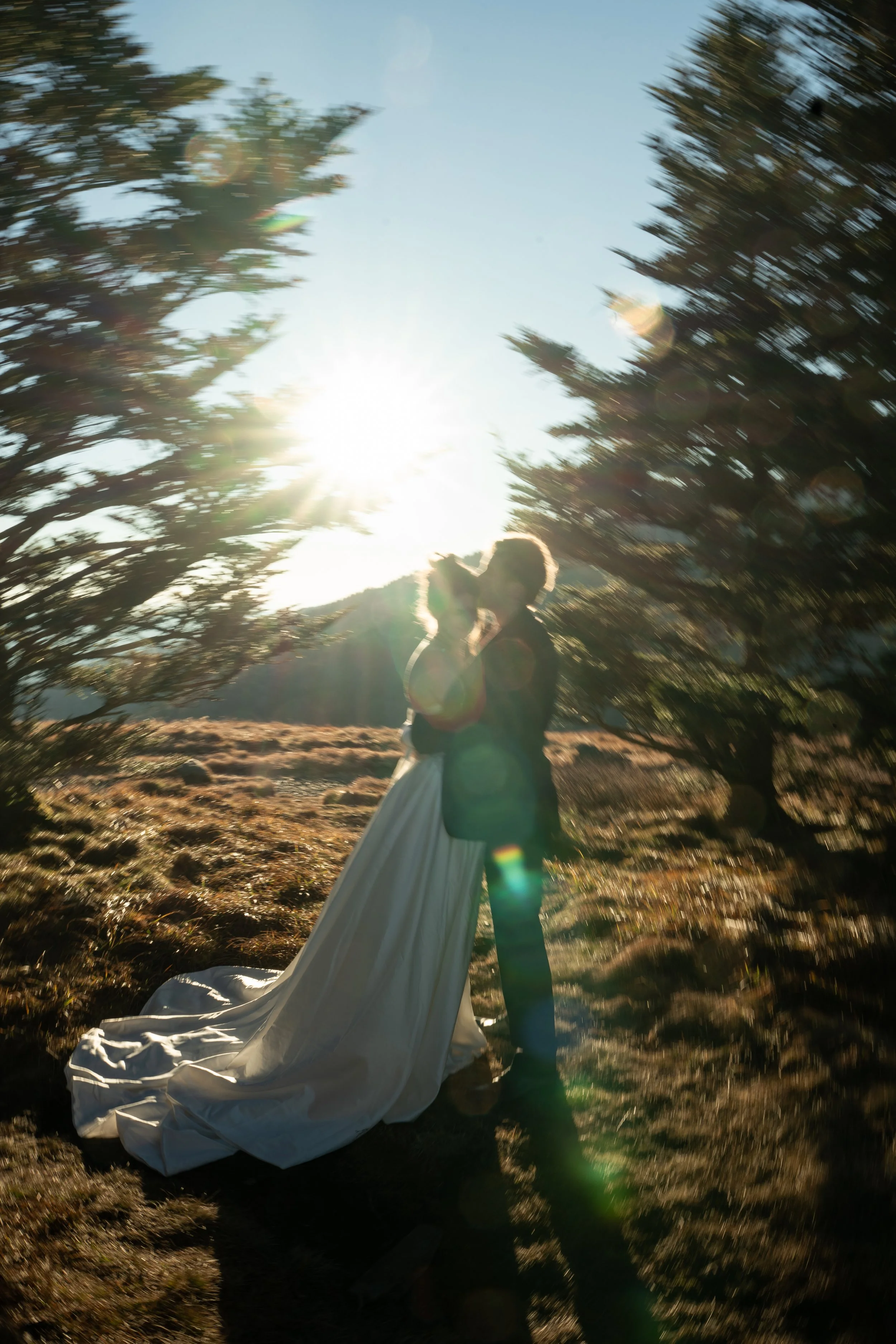 A couple, possibly newlyweds, standing outdoors surrounded by trees and grass during sunset, with the woman in a long white dress and the man in dark clothing, embracing and sharing a kiss.
