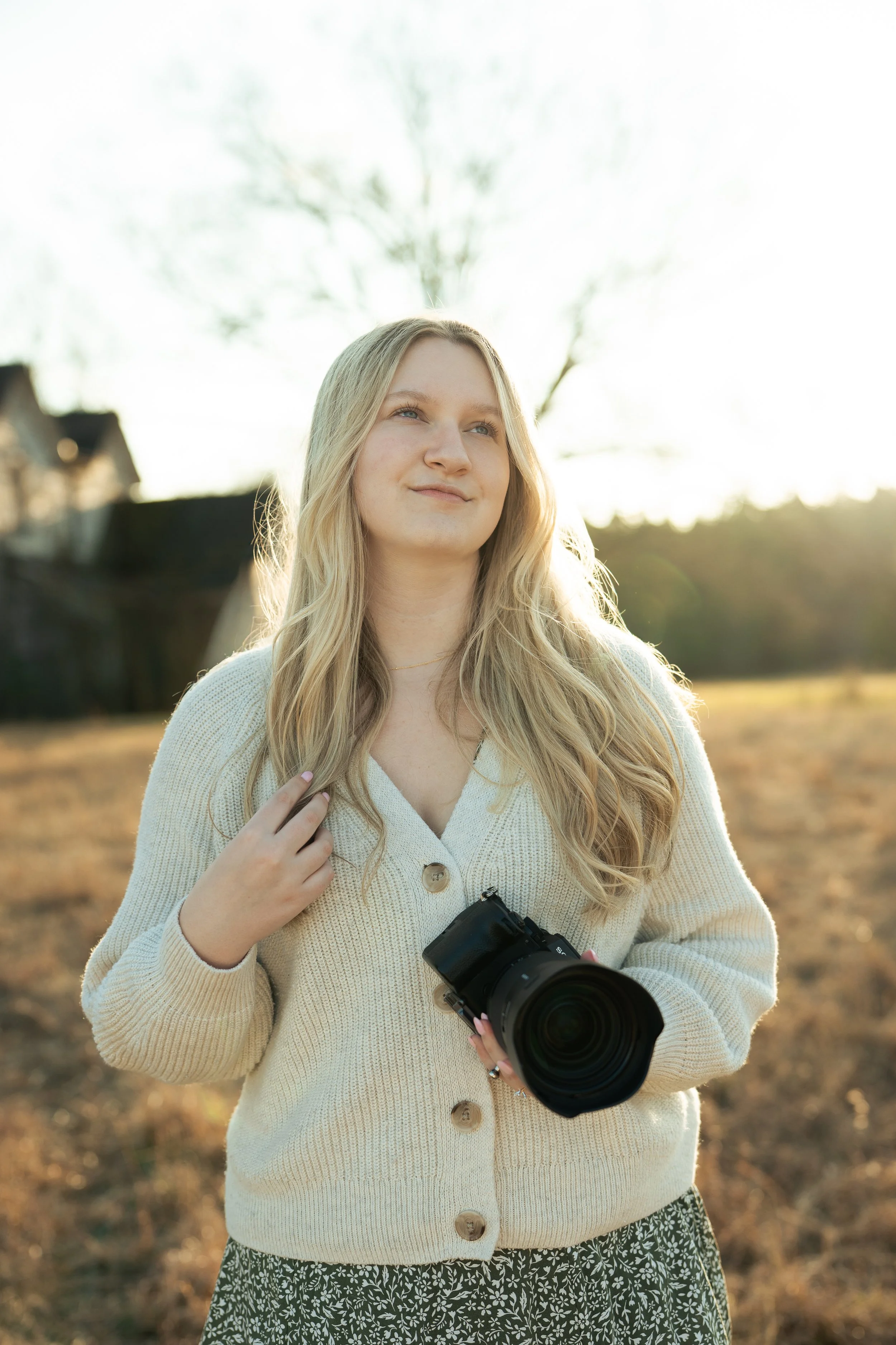 A young woman holding a camera outdoors in a field during sunset, with trees in the background.