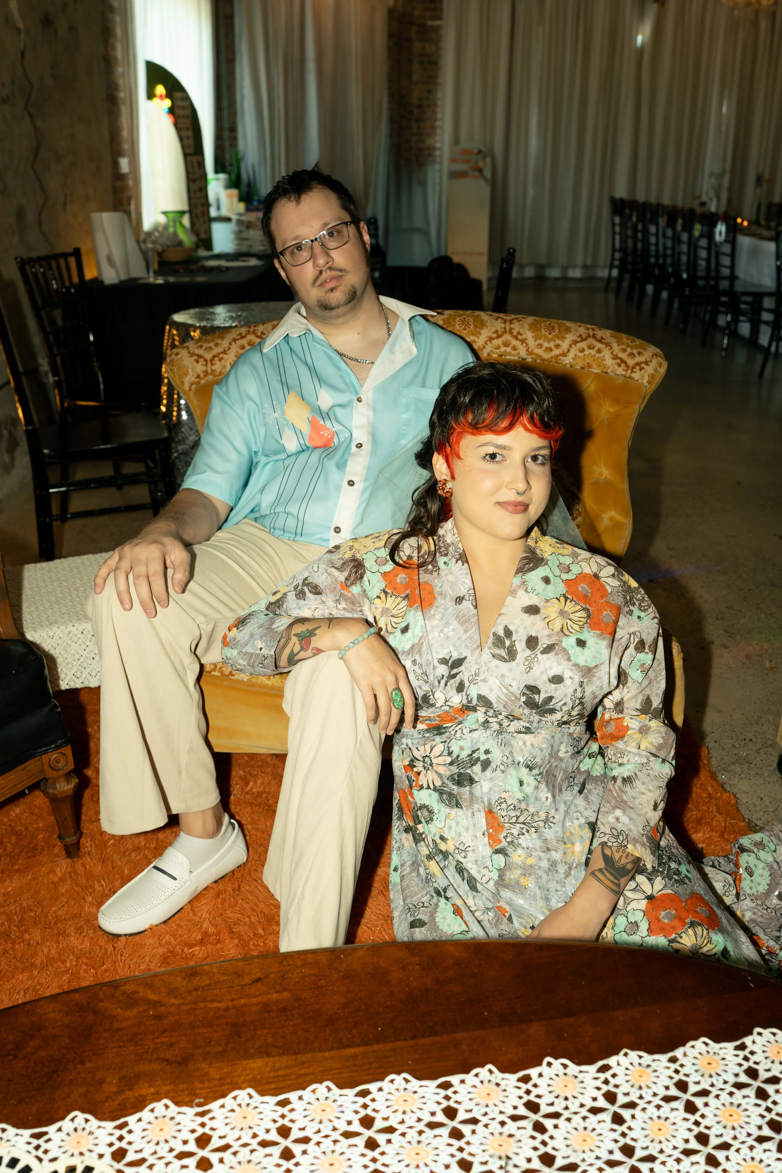A man and woman sitting together in a vintage-style living room or restaurant, with the man behind the woman. The man has short dark hair, glasses, and facial hair, and wearing a light blue retro shirt and beige pants. The woman has short dark hair with red highlights, multiple facial piercings, tattoos, and is wearing a floral-patterned shirt. There are chairs and tables in the background, and a lace tablecloth on a table in the foreground.