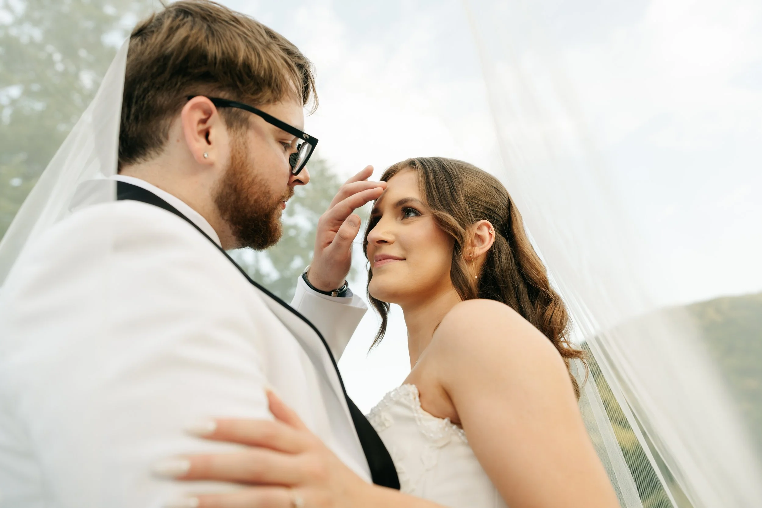 A groom and bride during a wedding ceremony, with the groom touching the bride's forehead while she gazes at him, both dressed in wedding attire outdoors with natural lighting.