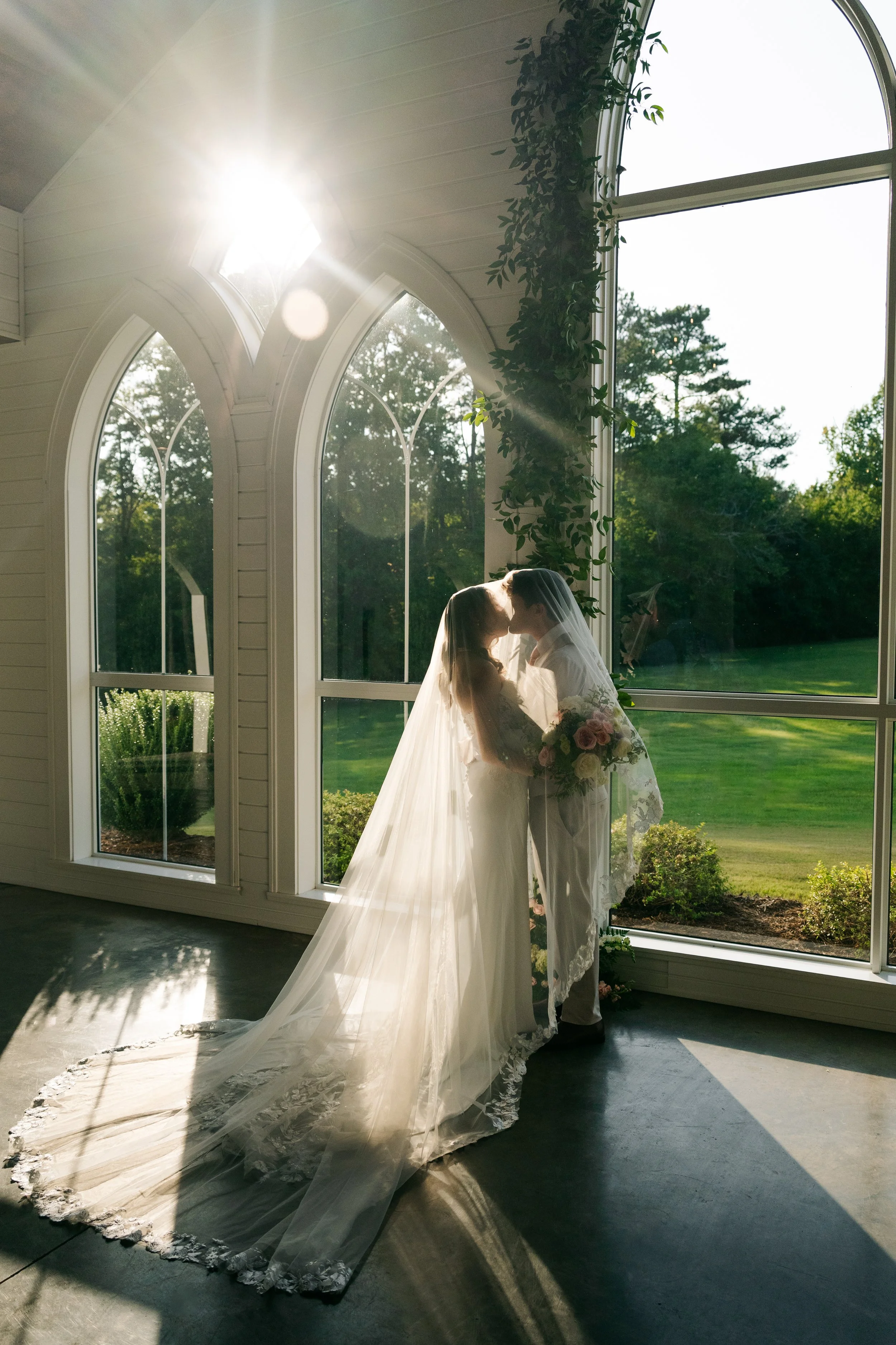 A bride and groom kissing inside a room with large arched windows, sunlight streaming in, and a view of trees and greenery outside. The bride is wearing a white wedding gown with a long train and veil, holding a bouquet of flowers.