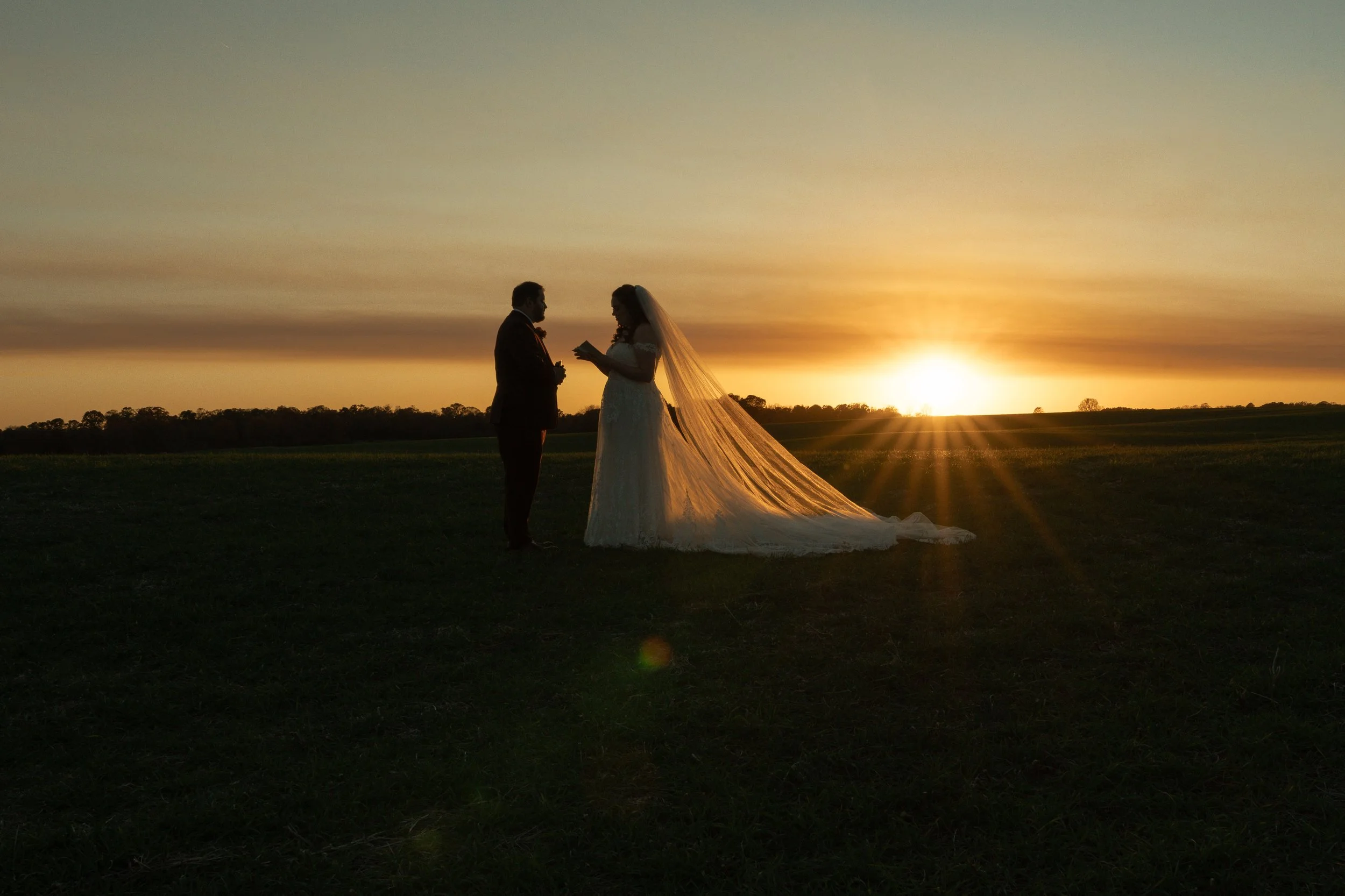 Silhouetted bride and groom standing on grass field during sunset, with bride reading vows or a letter, and sun rays illuminating their wedding dress train.