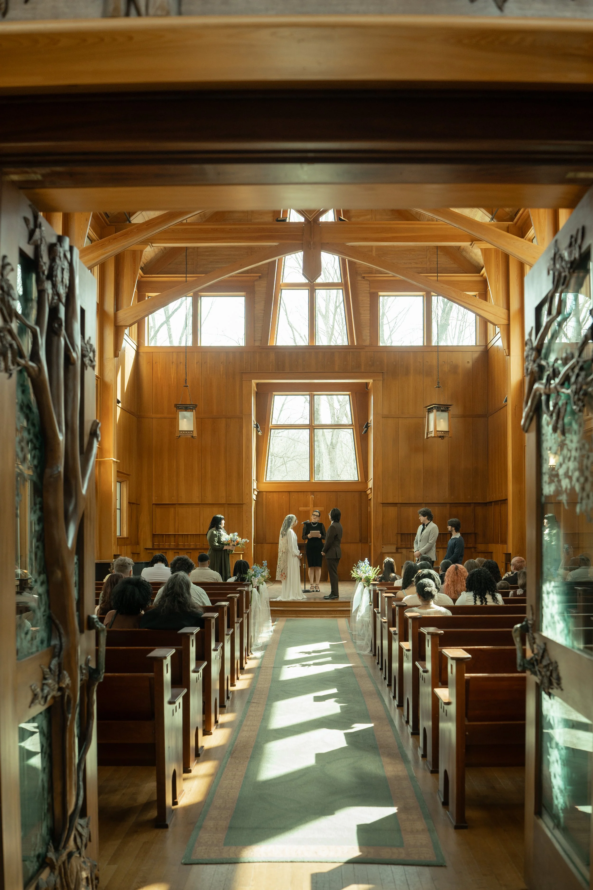 A wedding ceremony taking place inside a wooden church with large windows and natural light. The bride and groom stand at the altar with officiants, surrounded by guests seated in pews.