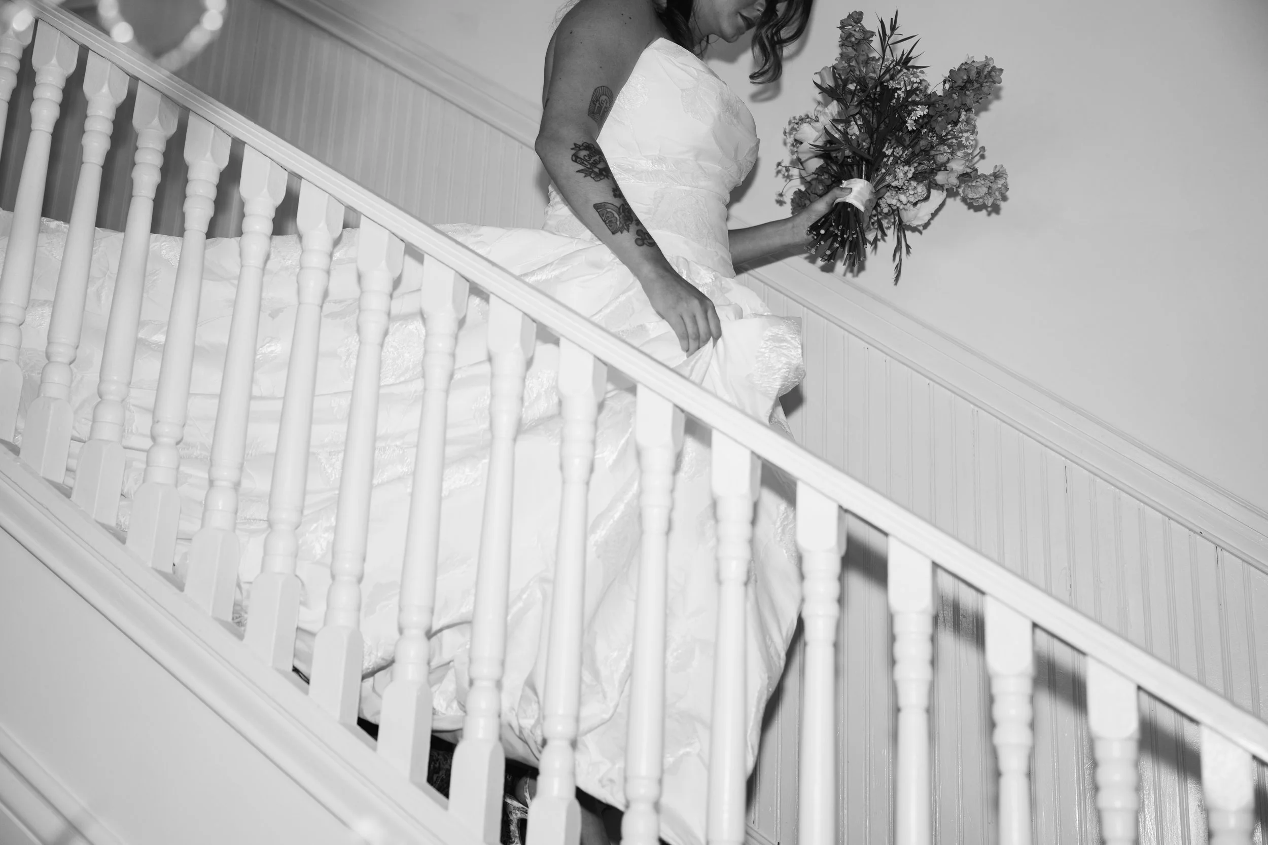 A woman in a wedding dress holding a bouquet of flowers, descending a staircase with wooden spindles, in black and white.