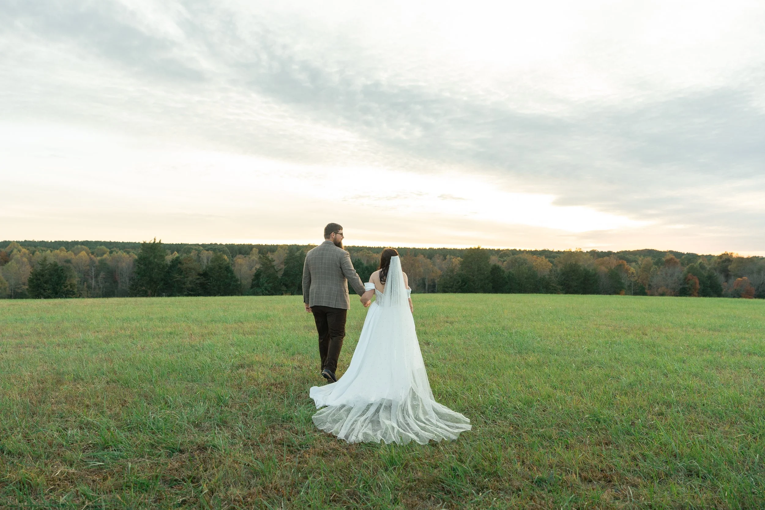 Couple walking hand-in-hand across a grassy field during sunset, with a line of trees in the background.