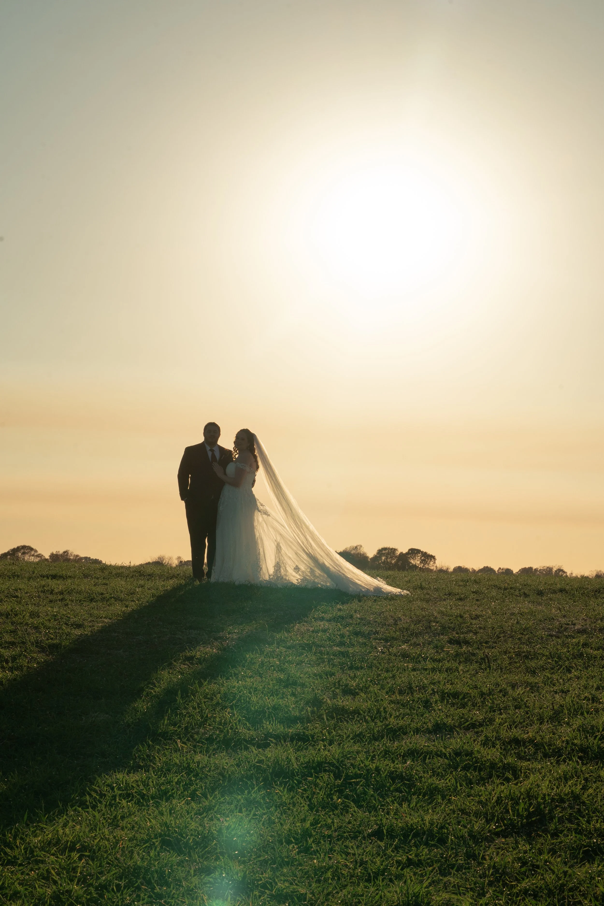 A couple in wedding attire standing on a grassy hill during sunset, with the bride in a long veil and wedding gown and the groom in a suit. The sun is bright and low in the sky, creating a silhouette effect and a long shadow.