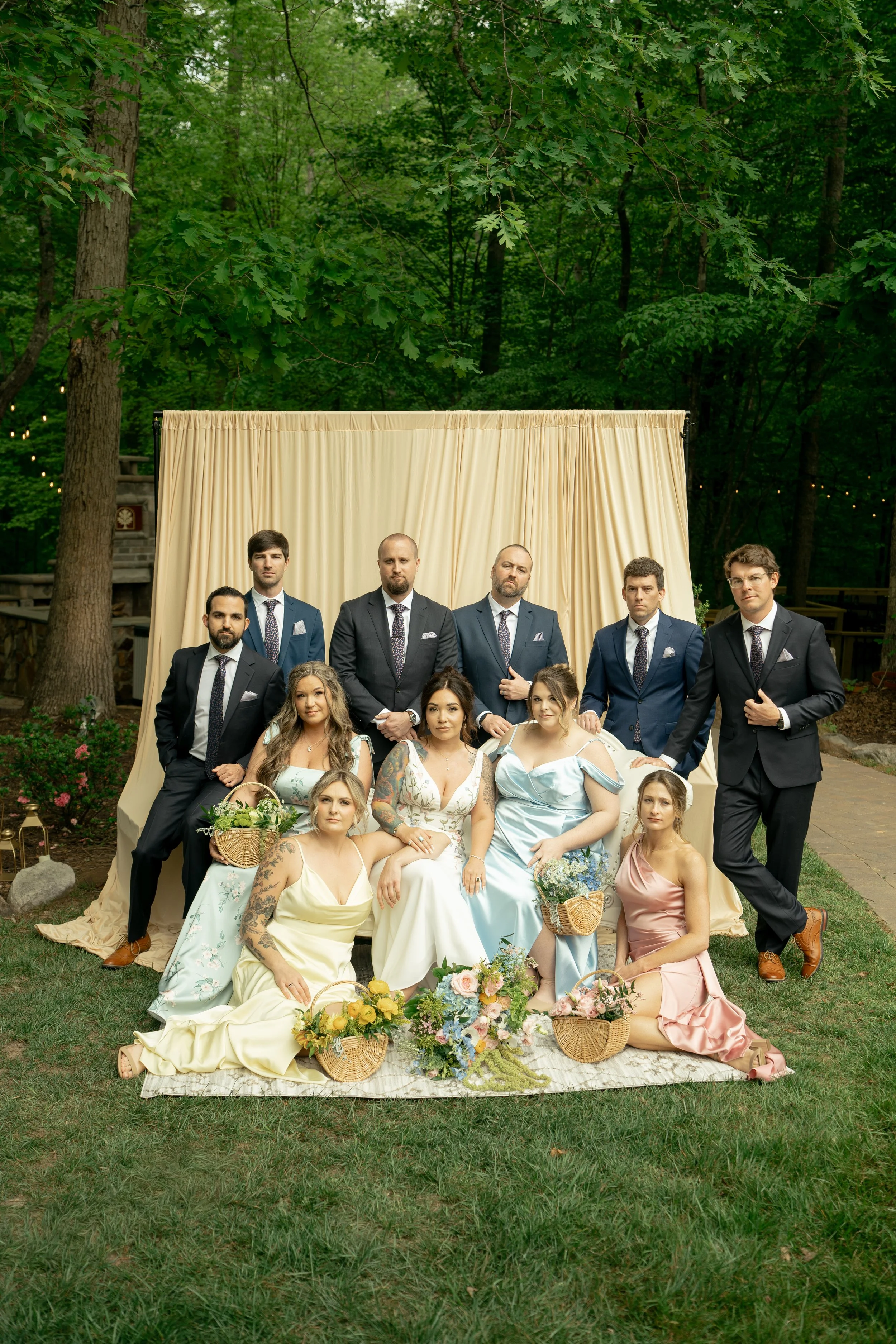 A group of people dressed in formal attire posing outdoors in front of a cream-colored curtain backdrop, surrounded by trees and greenery. The group includes men in suits and women in dresses, with floral arrangements and wicker baskets with flowers placed at their feet.