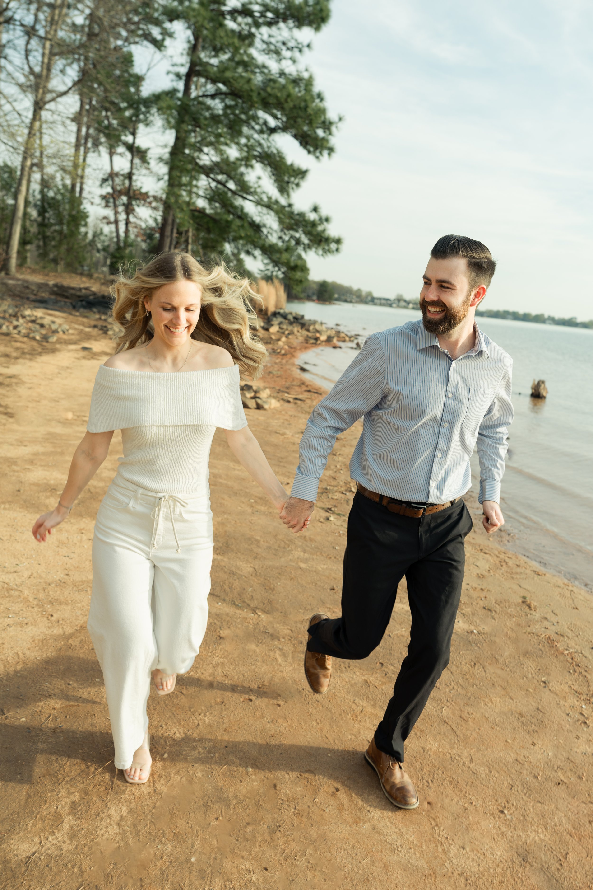A couple holding hands and running on a beach near a body of water, with trees in the background.