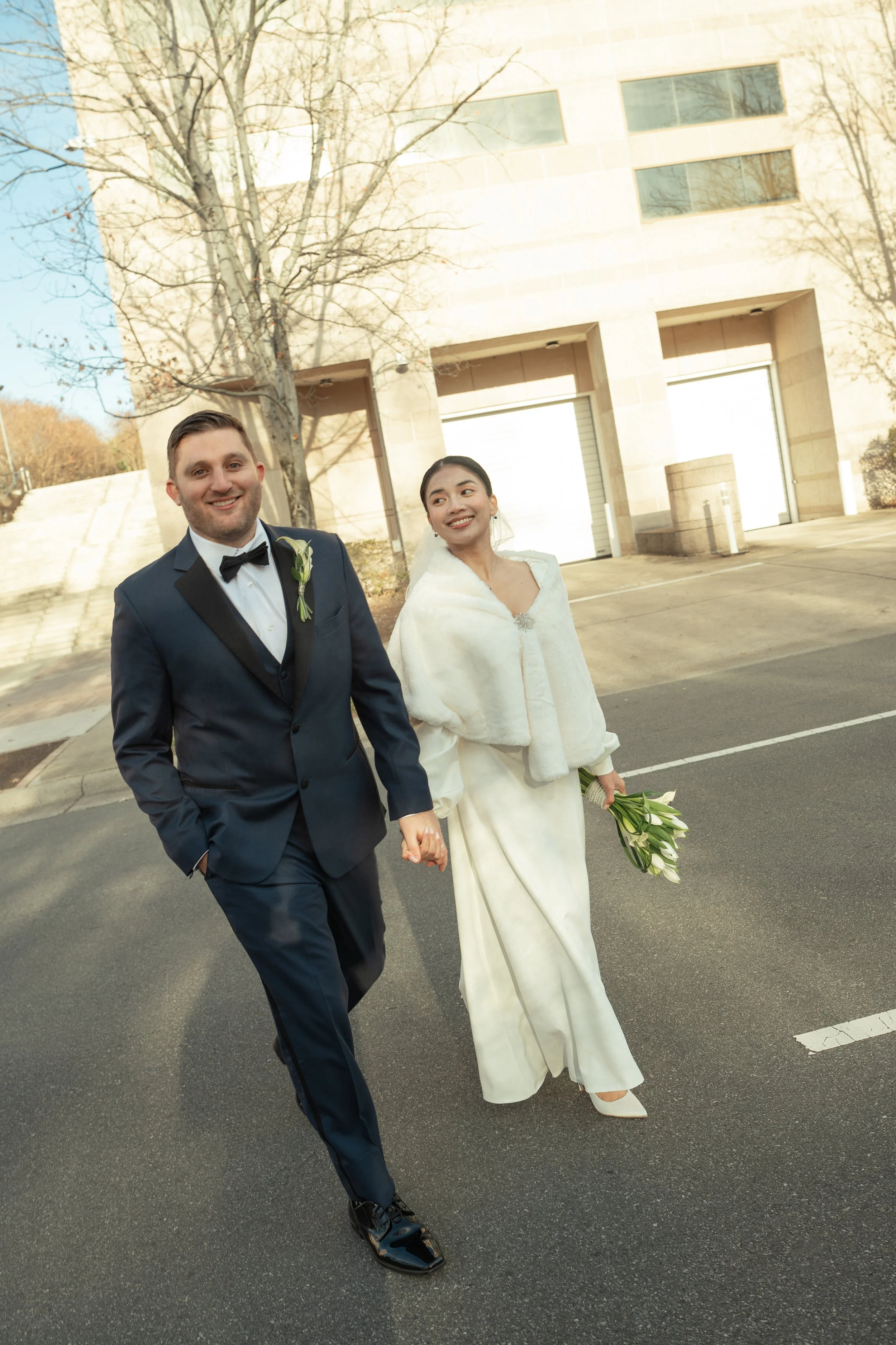 A newlywed couple walking hand in hand outdoors, the groom in a tuxedo and the bride in a white gown with a white shawl, holding a bouquet of white flowers, with a modern building and leafless trees in the background.