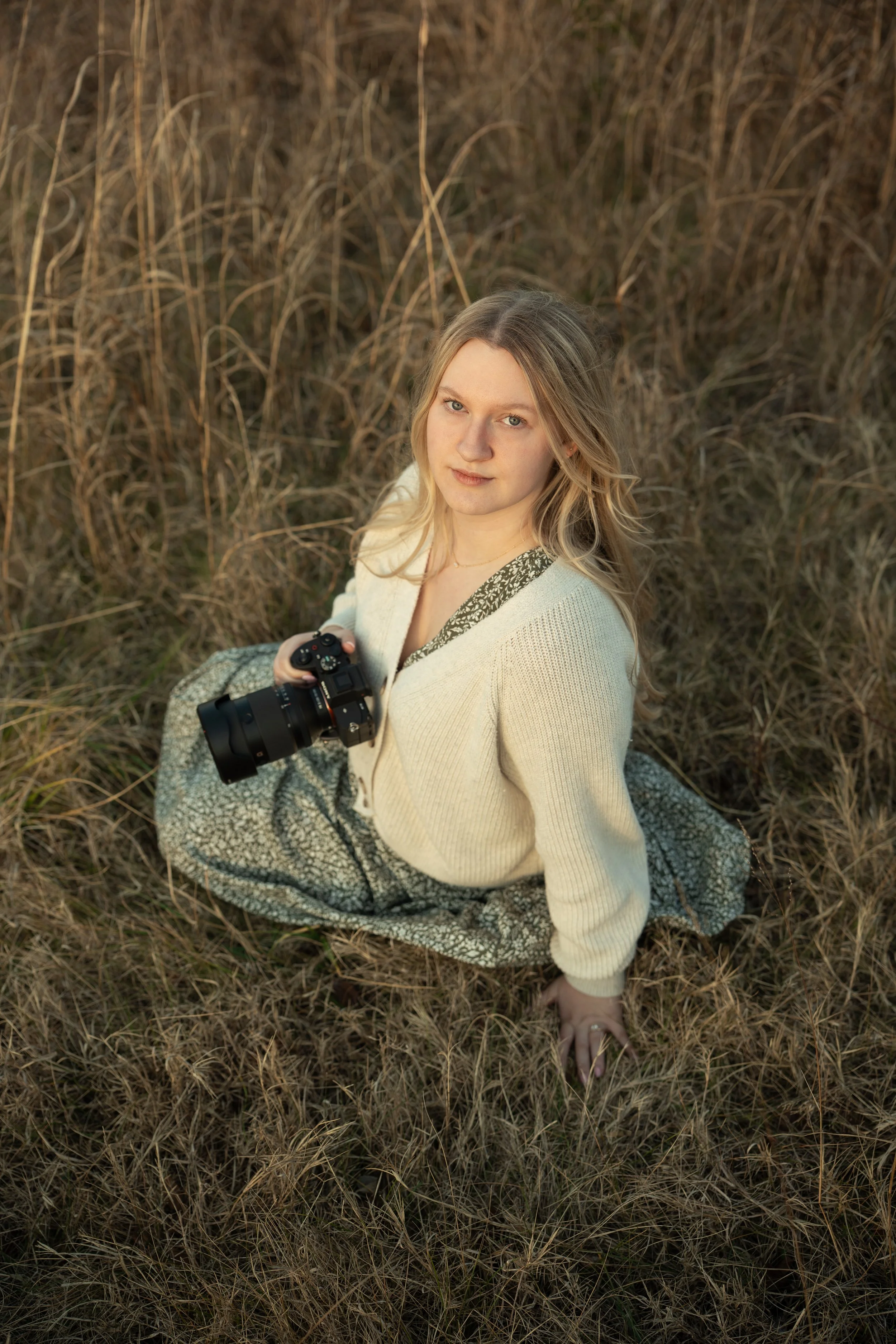 A young woman with blonde hair sitting on the ground in a field of tall grass, holding a camera, looking up at the camera with a slight smile.