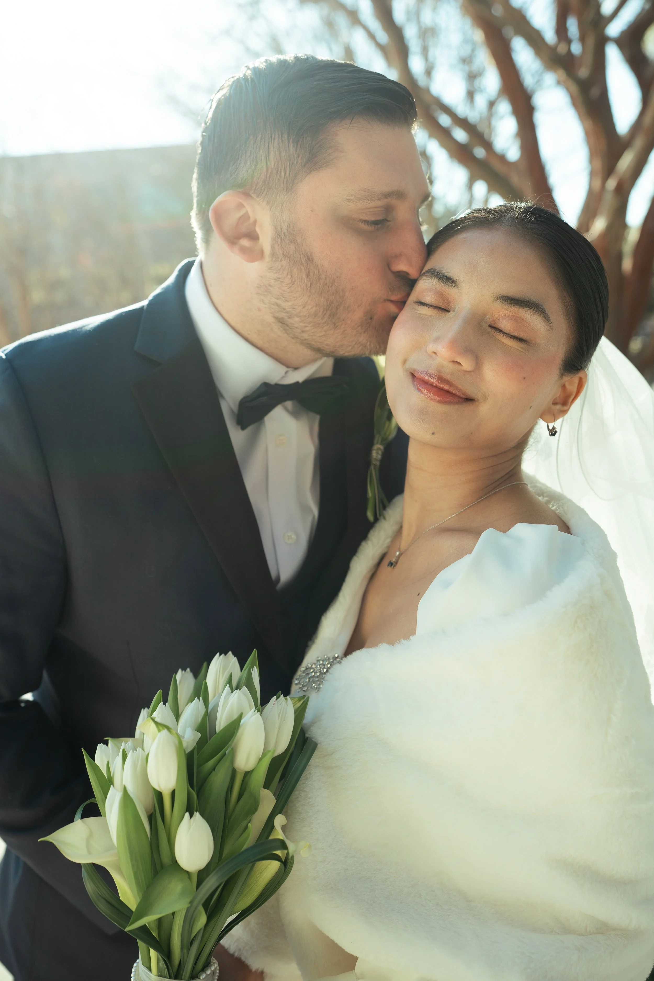 A bride and groom on their wedding day, the groom kissing the bride's forehead. The bride is holding a bouquet of white tulips and is wearing a white dress with a fur shawl. The groom is in a tuxedo with a bow tie. They are outside with sunlight shining through the trees.