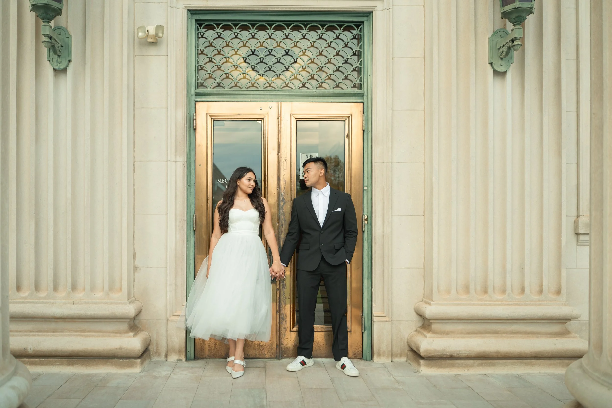 A couple holding hands in wedding attire standing in front of a vintage green and gold door on a stone building.