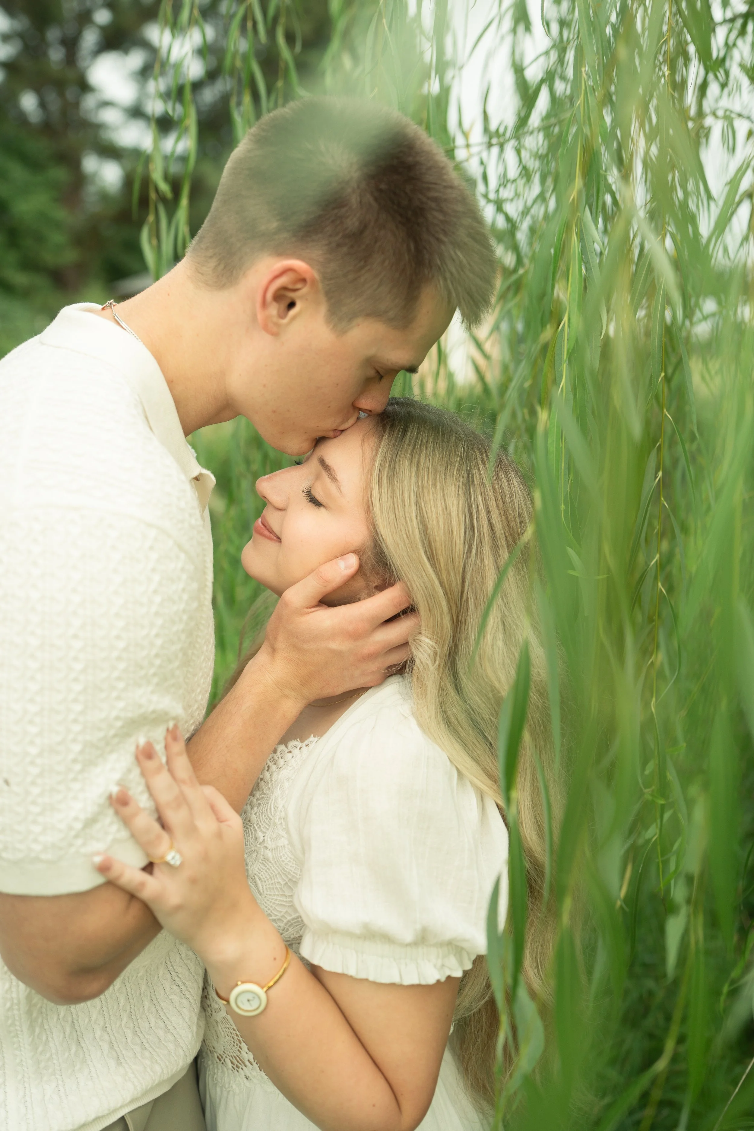 A young couple in white clothing embracing outdoors among green plants, with the man kissing the woman on her forehead and the woman smiling with her eyes closed.