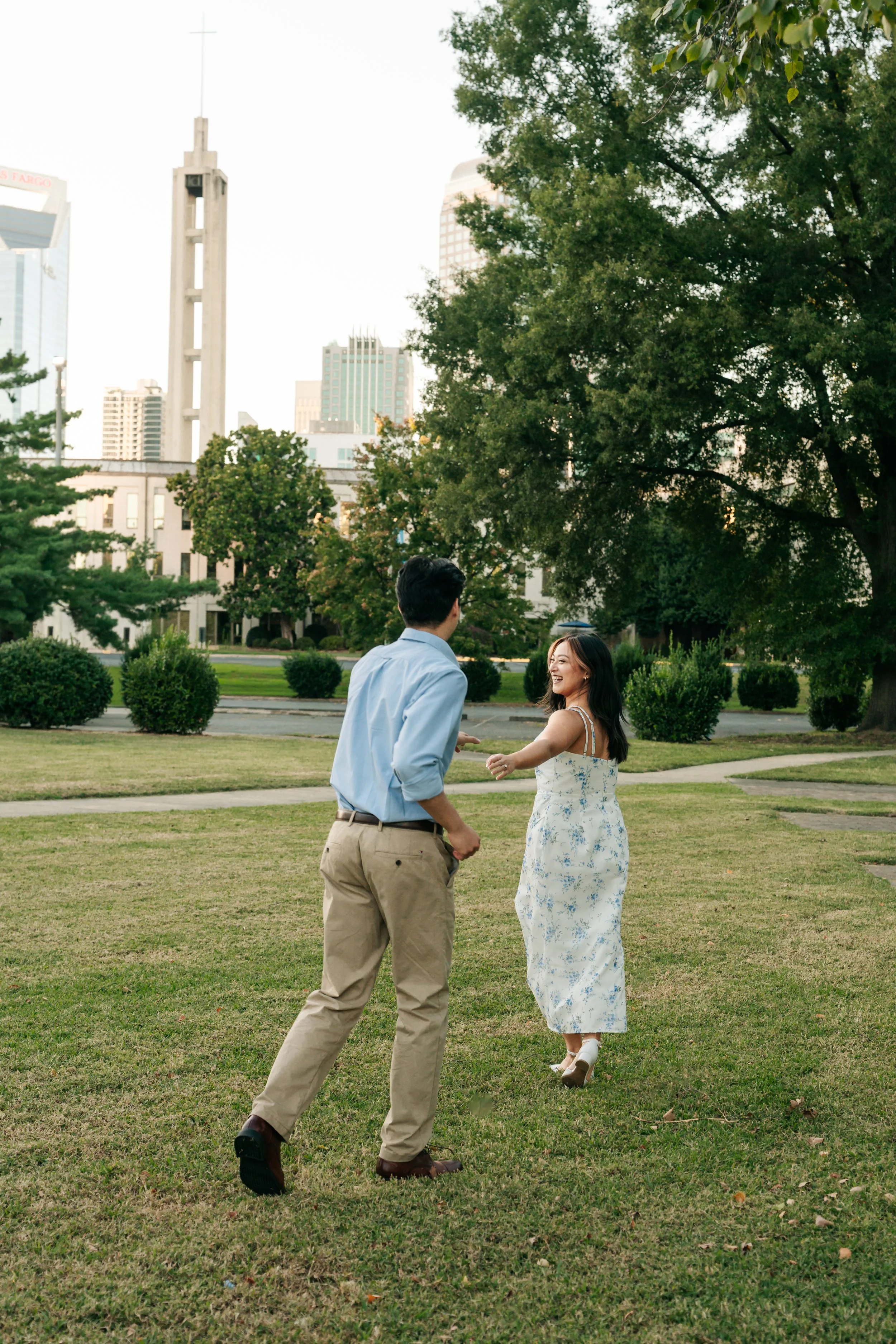 A young man and woman are dancing outside in a park with city skyscrapers in the background. The woman is wearing a white floral dress and high heels, and the man is wearing a light blue shirt and beige pants. They are smiling and holding hands, enjoying a moment together.