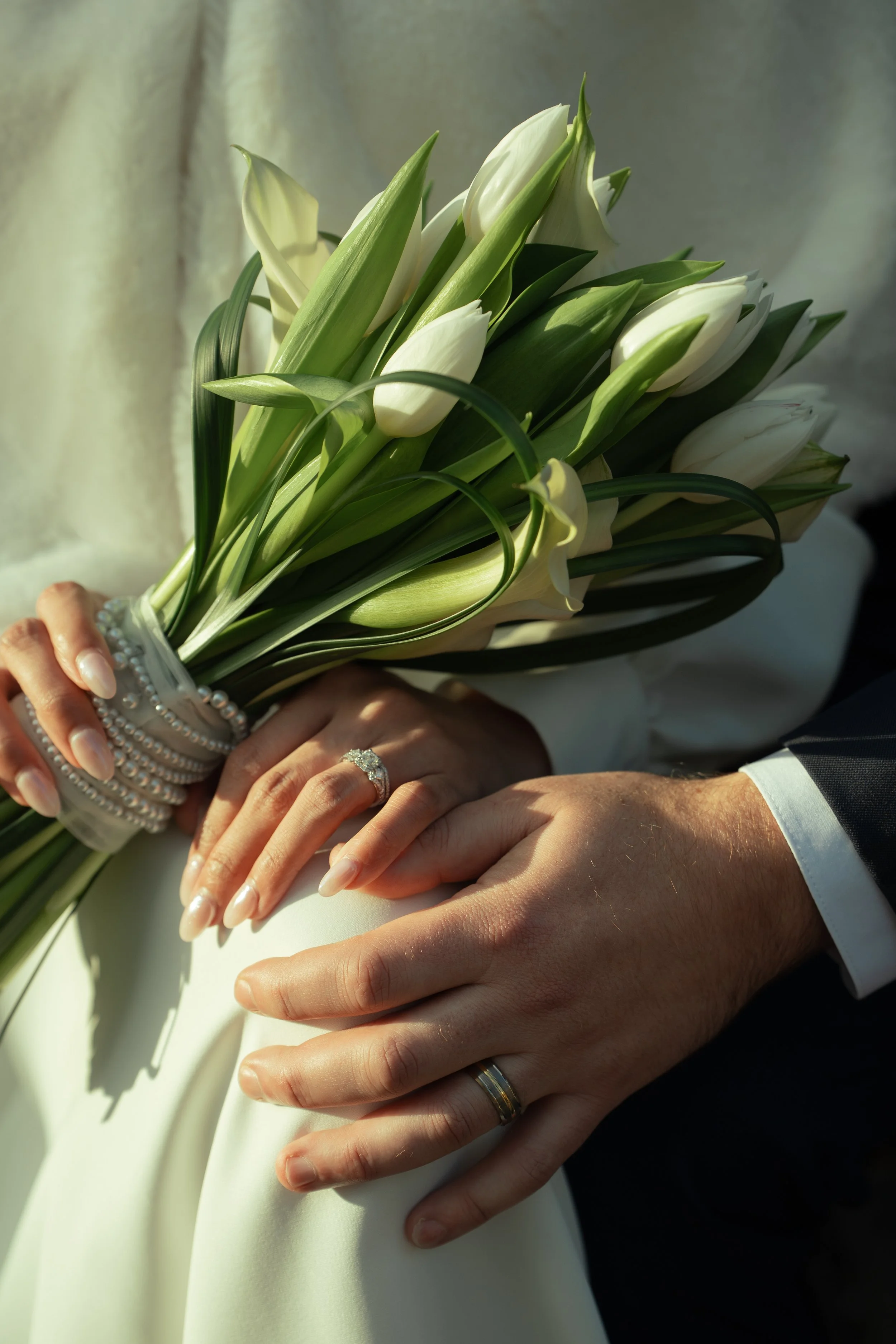 Close-up of a bride and groom's hands. The bride holds a bouquet of white calla lilies and wears a diamond ring and pearl bracelets. The groom's hand, with a wedding band, rests on the bride's dress.
