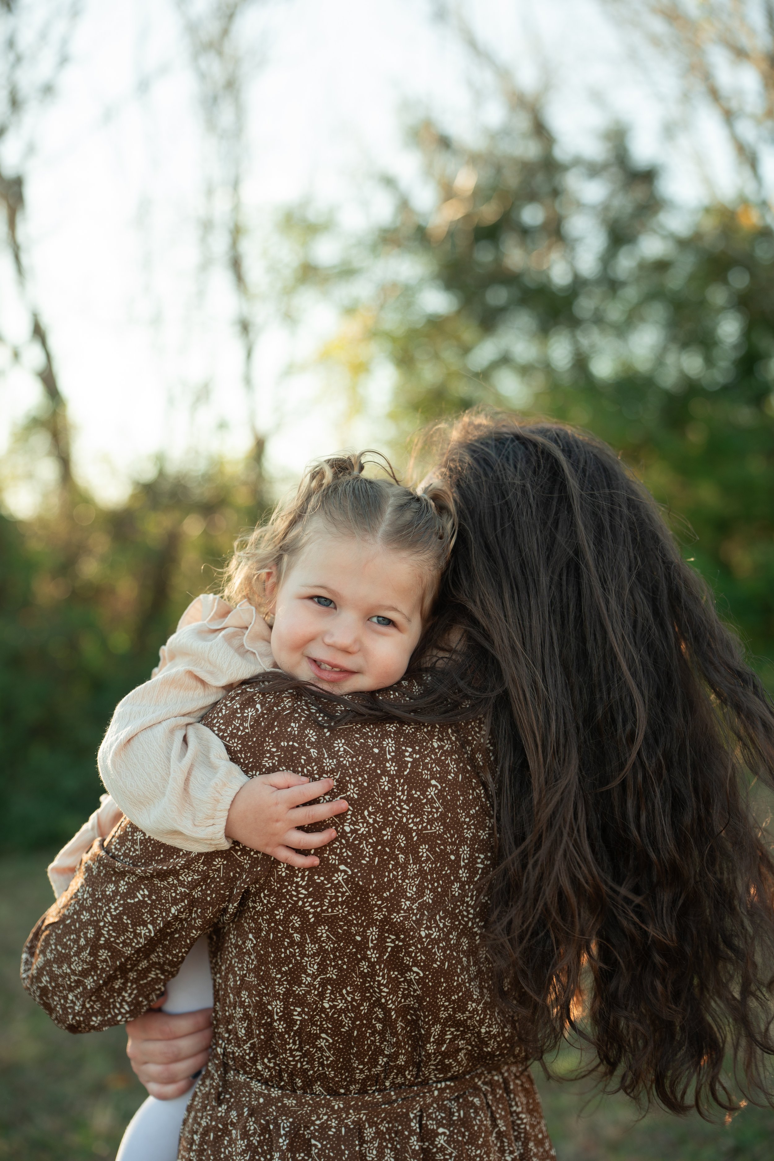 A young girl is being carried piggyback by a woman outdoors, with trees and sunlight in the background.