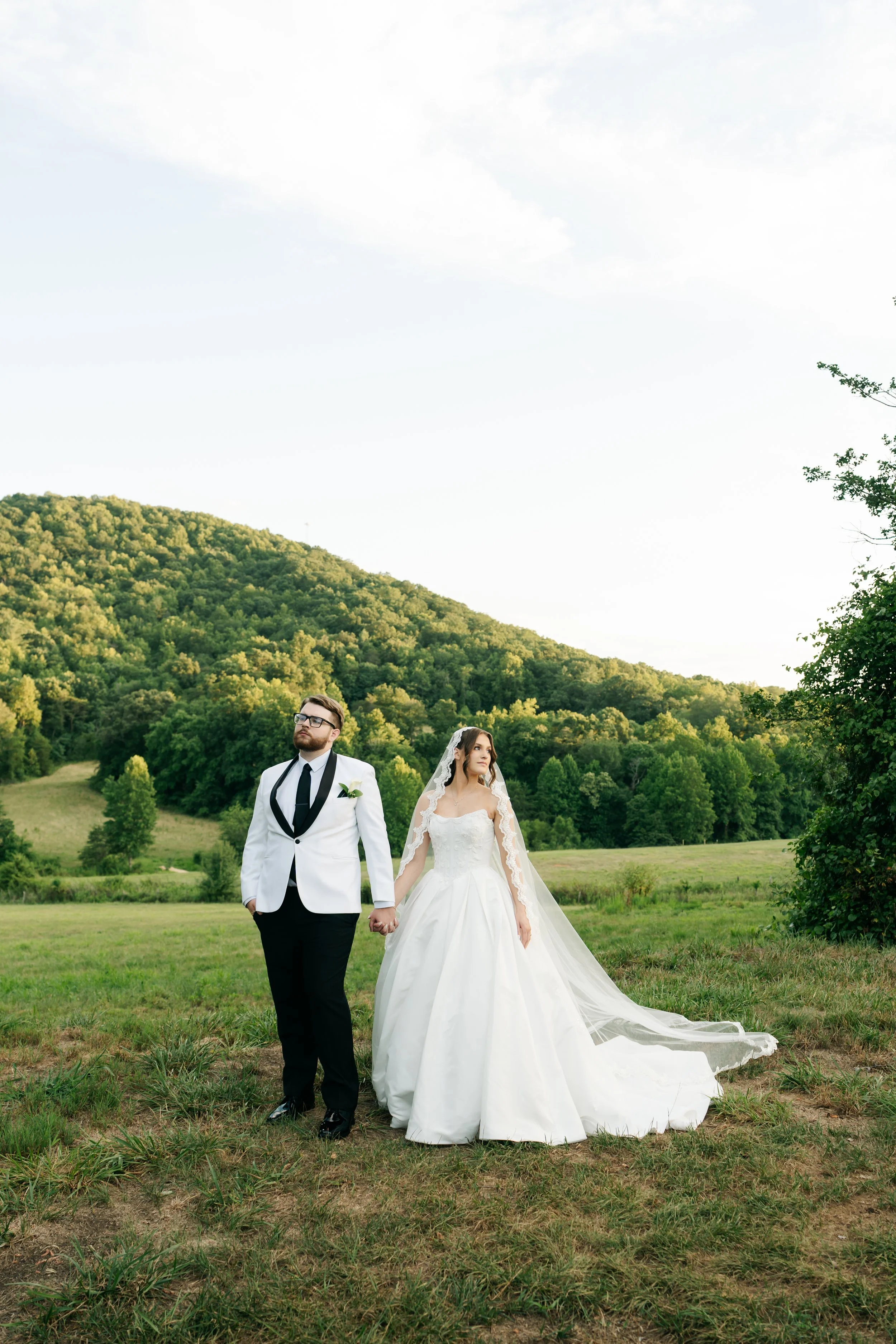 A bride and groom holding hands and walking outdoors in a lush green field with trees and a hill in the background during daylight.