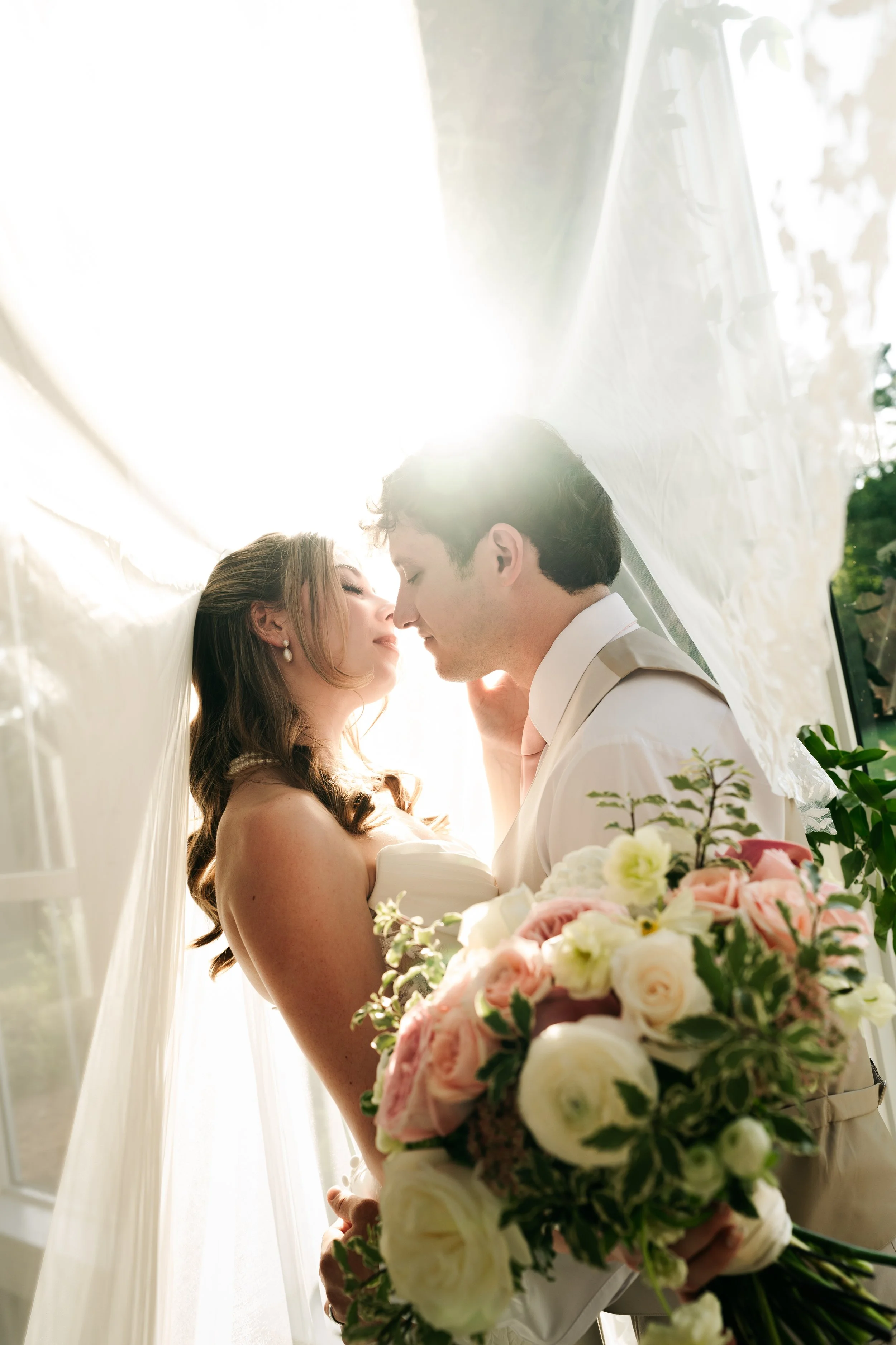 Bride and groom standing close under a floral canopy, smiling with foreheads touching, sunlight shining on them, bride holding a bouquet of pink and white roses and greenery.