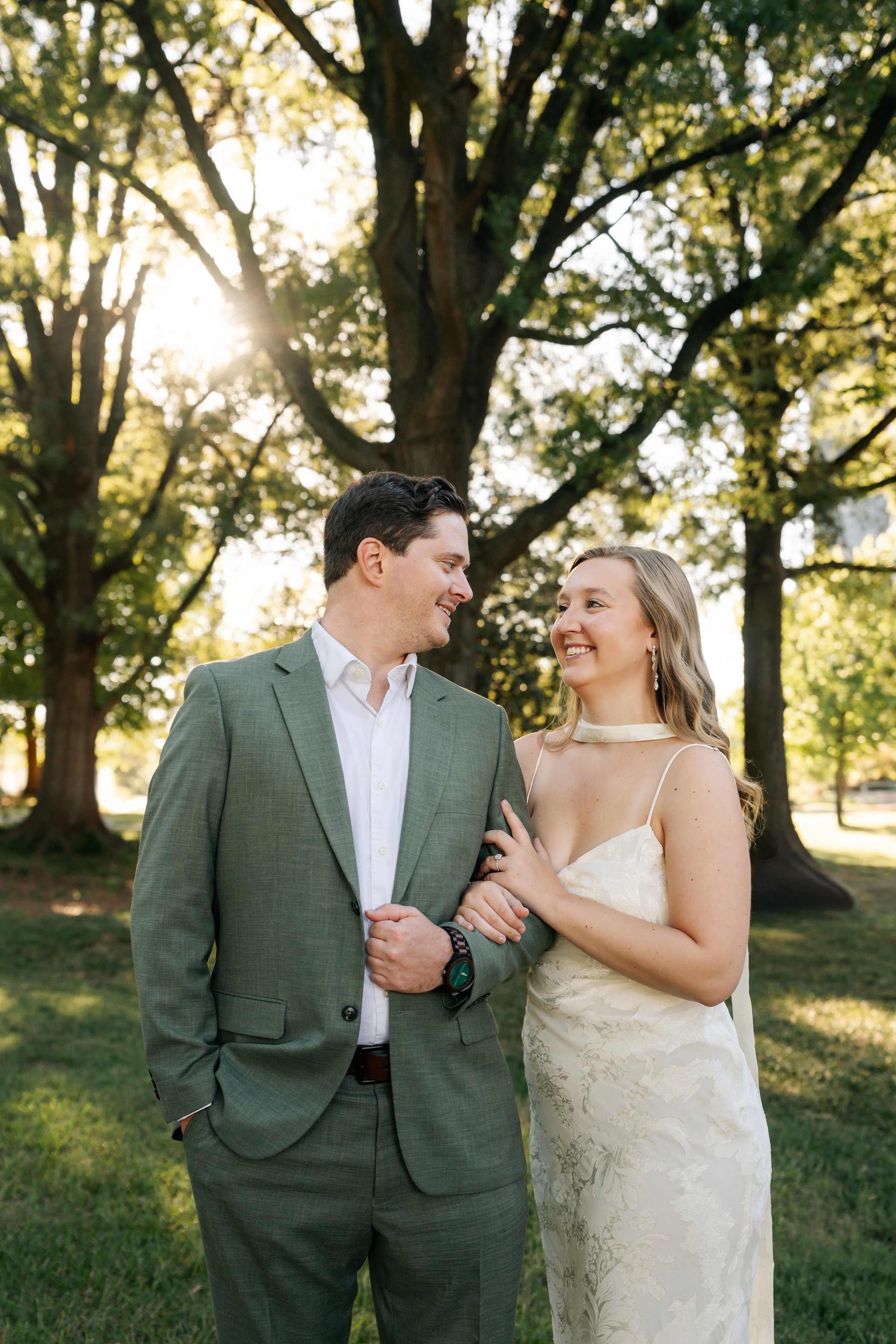 A couple in wedding attire standing close together outdoors beneath trees with sunlight filtering through the leaves, smiling at each other.
