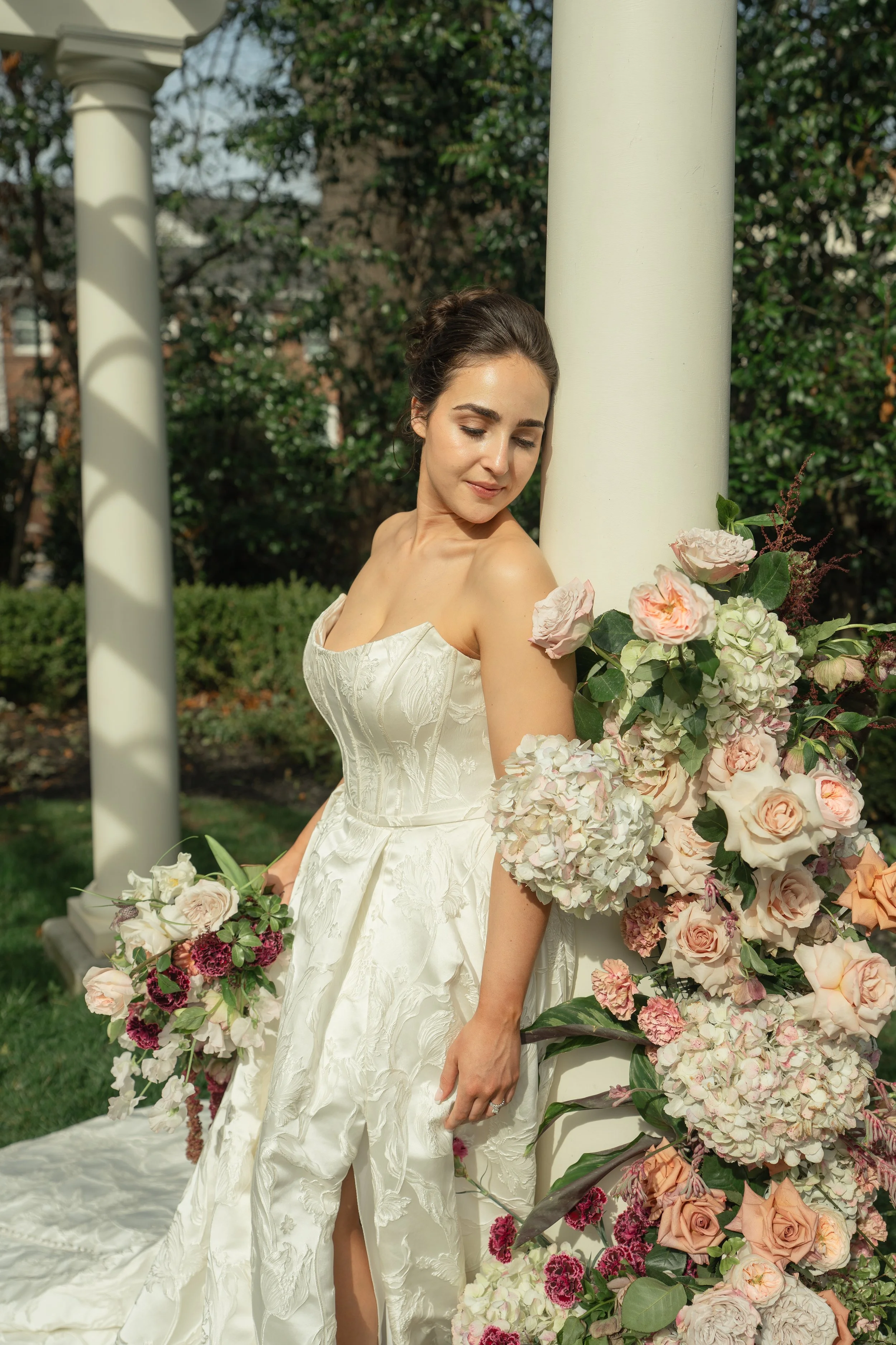 A woman in a bridal gown leaning against a white column surrounded by pink and white flowers outdoors.