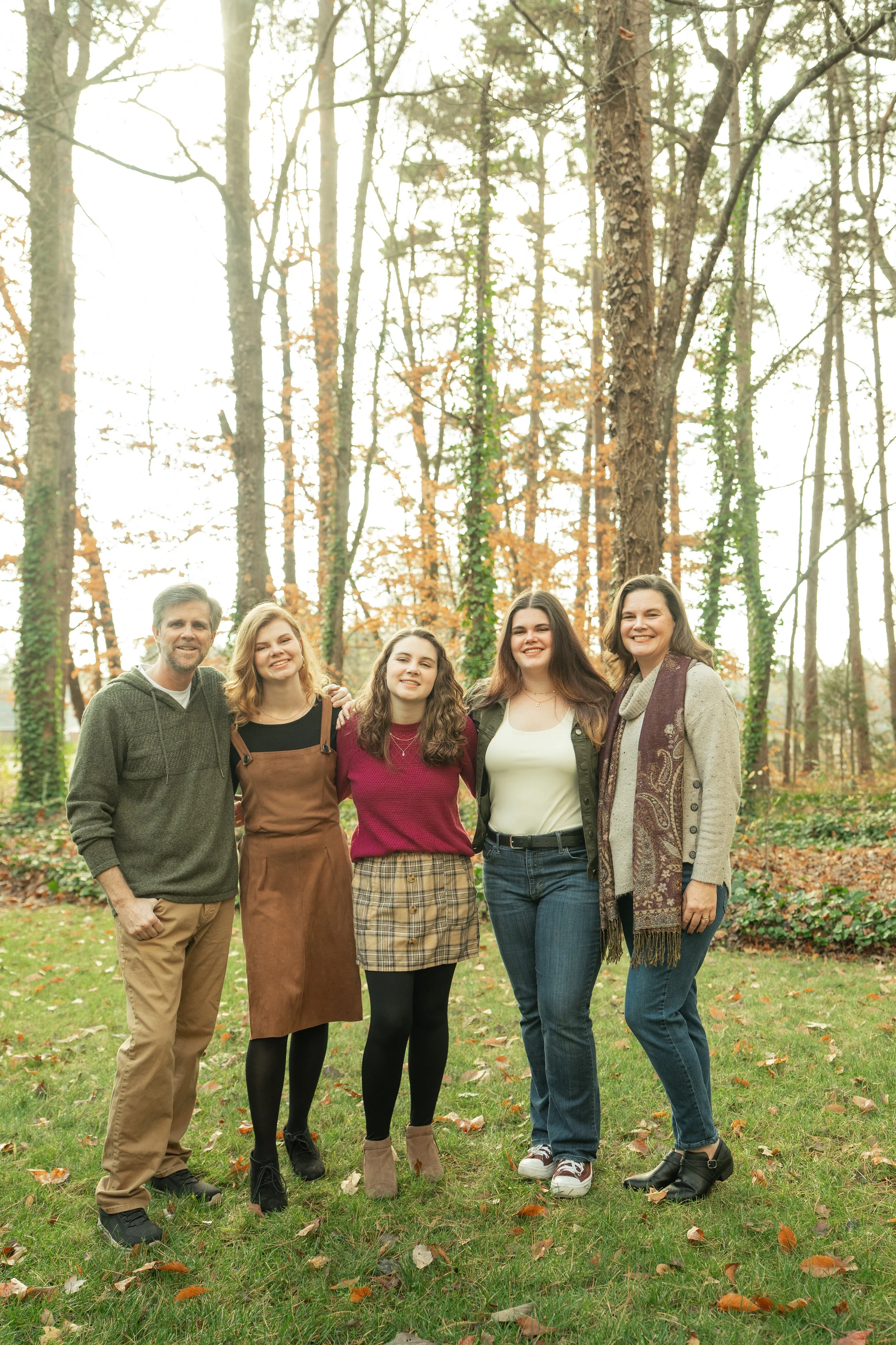A family of five standing outdoors on a grassy area with scattered autumn leaves, surrounded by trees with some orange and yellow foliage.