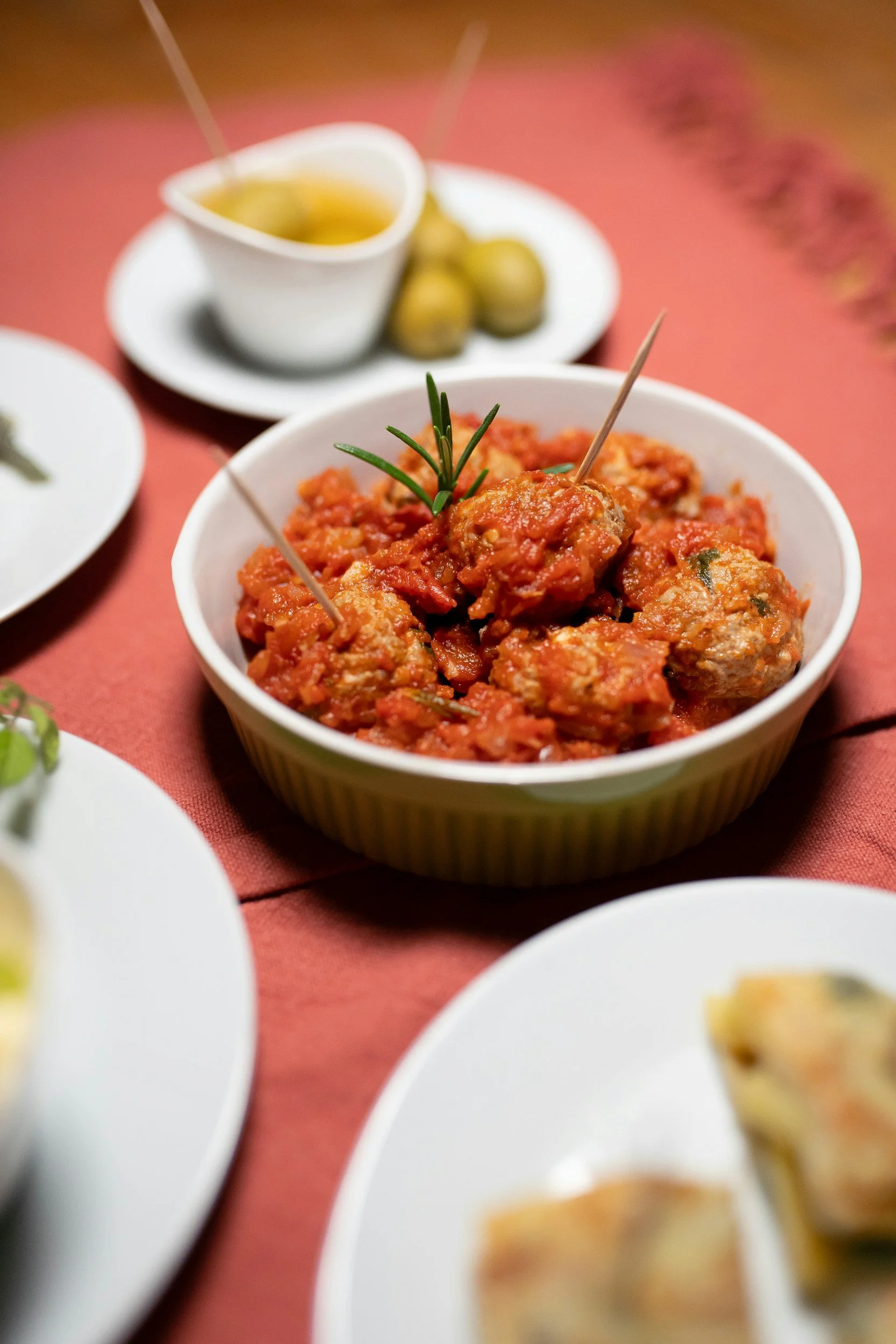 A bowl of meatballs in tomato sauce garnished with a sprig of rosemary, surrounded by other plates of food on a table.
