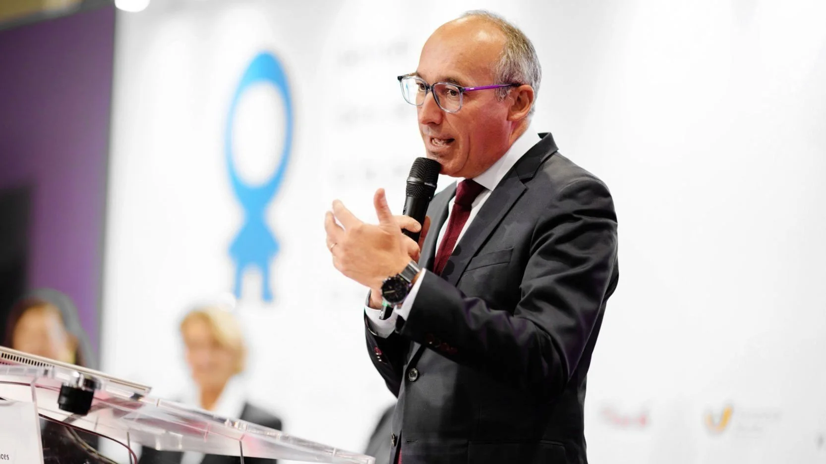 Allocution d’un intervenant en conférence, micro à la main, fond institutionnel et mise en scène corporate.