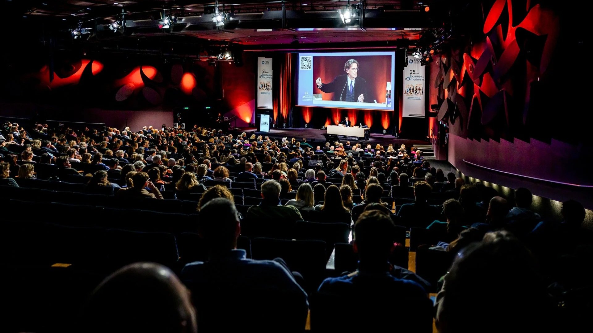 Conférence professionnelle devant un large public, scène éclairée et présentation projetée.