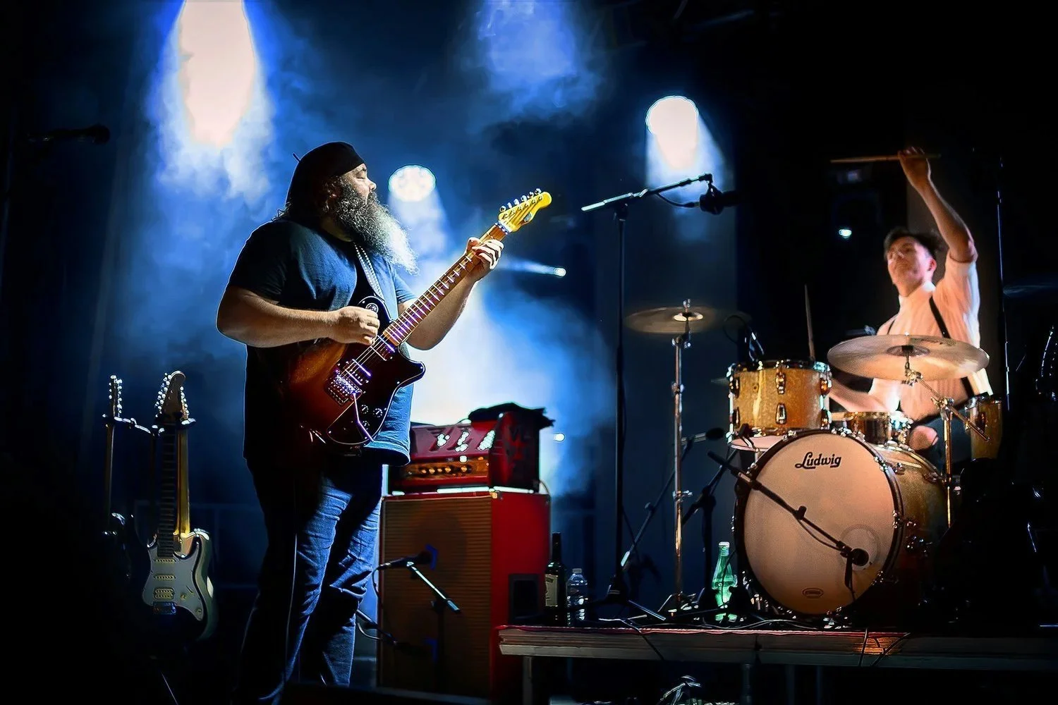Groupe de musique sur scène, interaction entre guitariste et batteur captée en photo live.