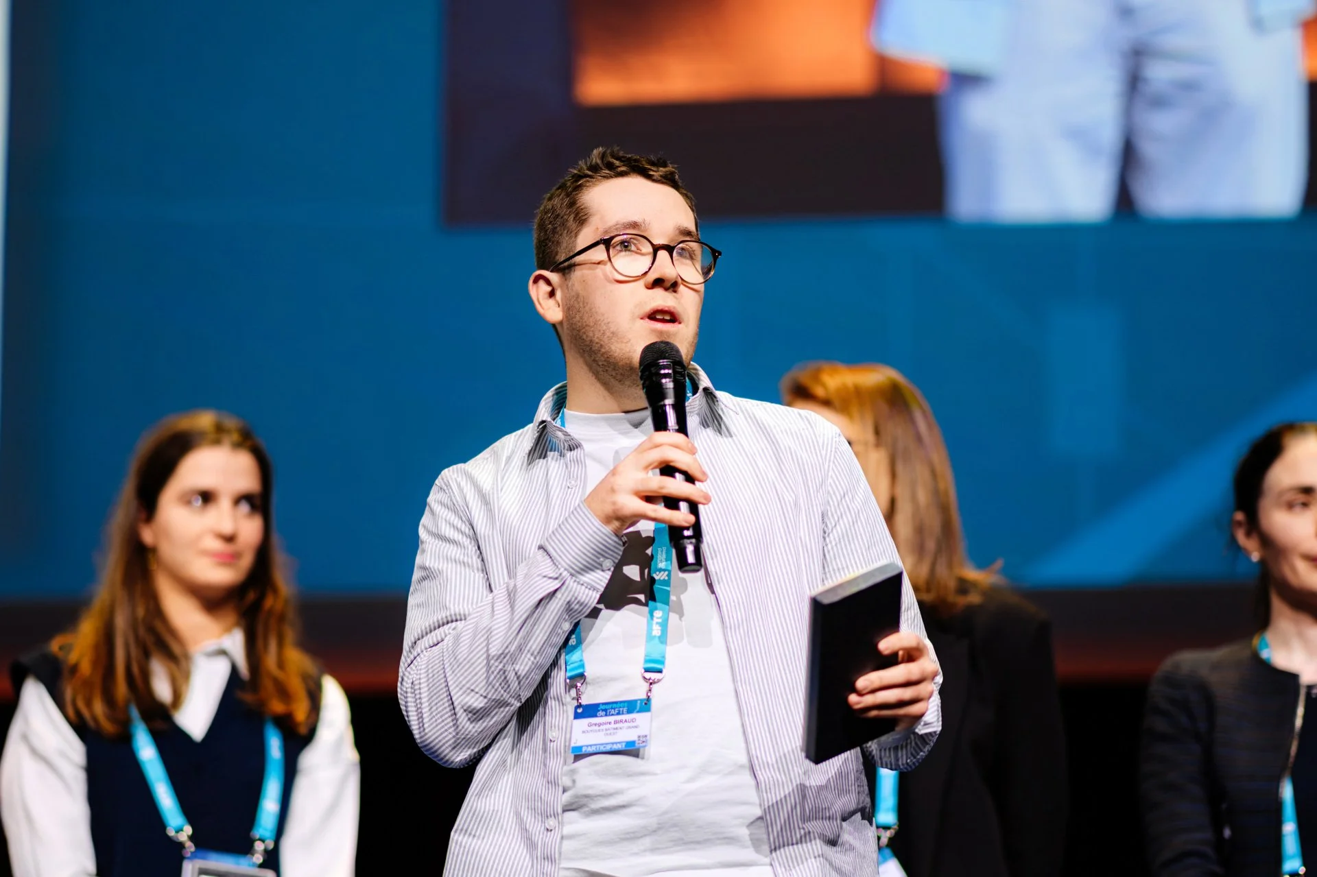 Portrait d’un intervenant des Journées AFTE 2025 prenant la parole sur scène avec un micro, devant un écran de conférence.