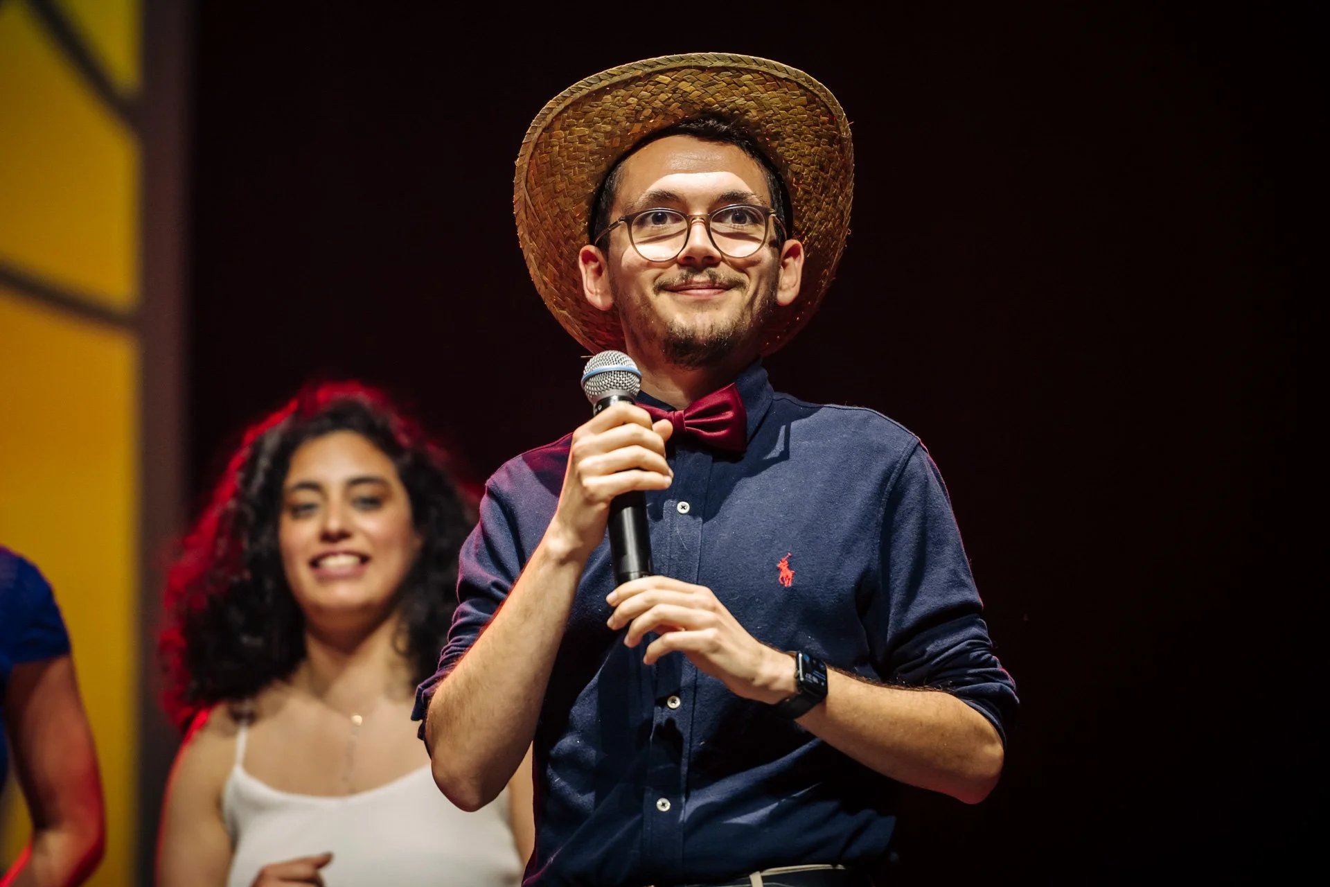 Portrait d’un étudiant du CBH Graduate School, micro à la main et chapeau de paille, prenant la parole devant le public du gala à Grenoble.