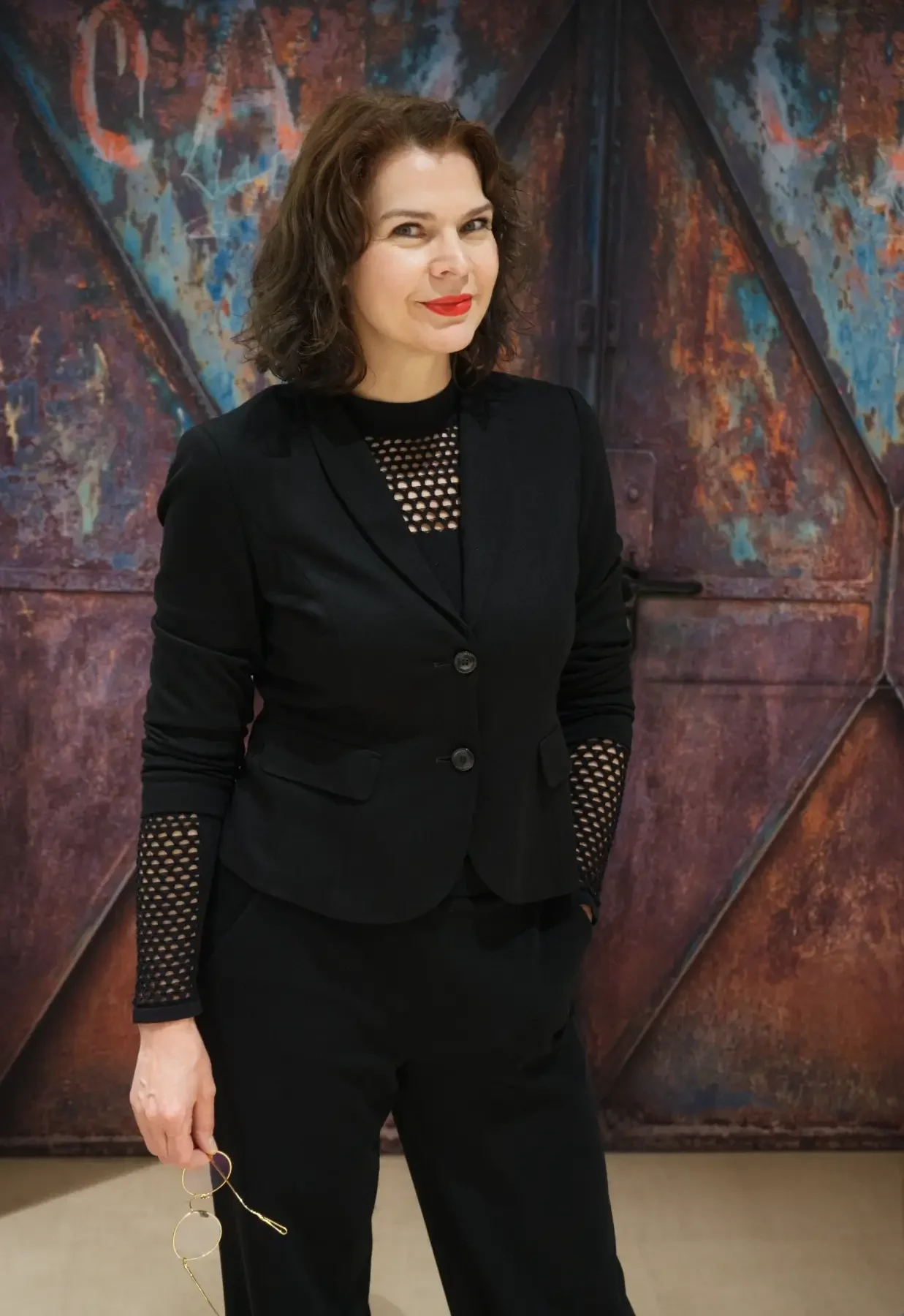 Alexandra Poser steht in einem schwarzen Anzug vor einer farbig gestalteten Wand und hält eine Brille in der Hand.