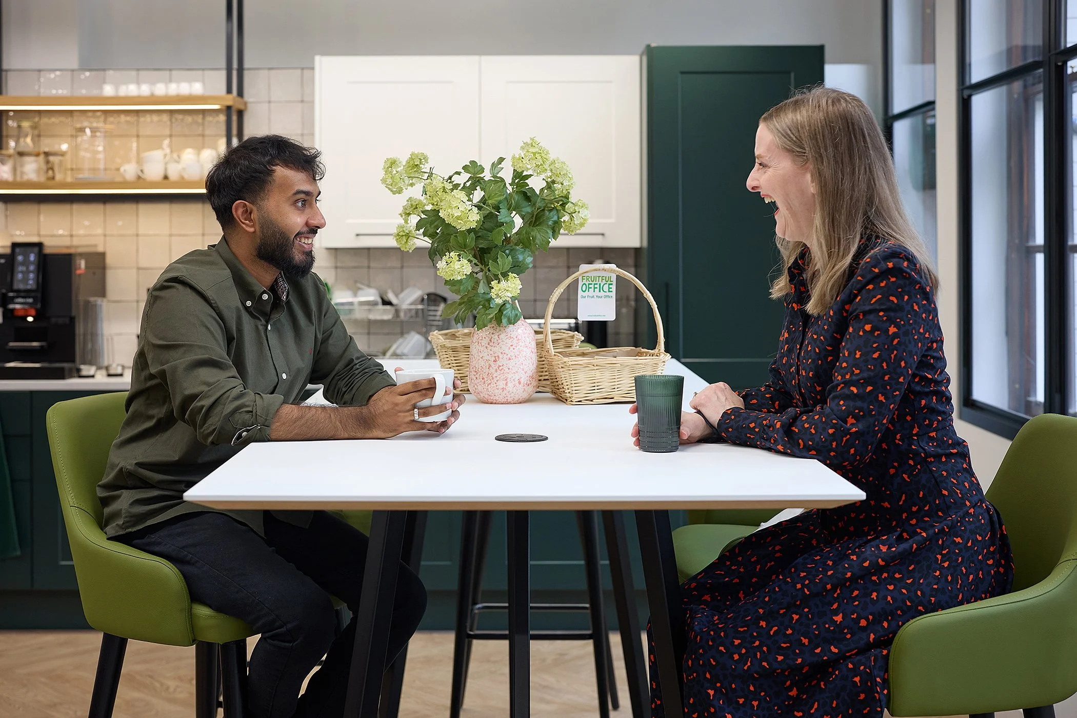 Two people, man and woman, sitting at a white table in a kitchen, smiling and talking, each holding a cup, with a plant and baskets on the table.