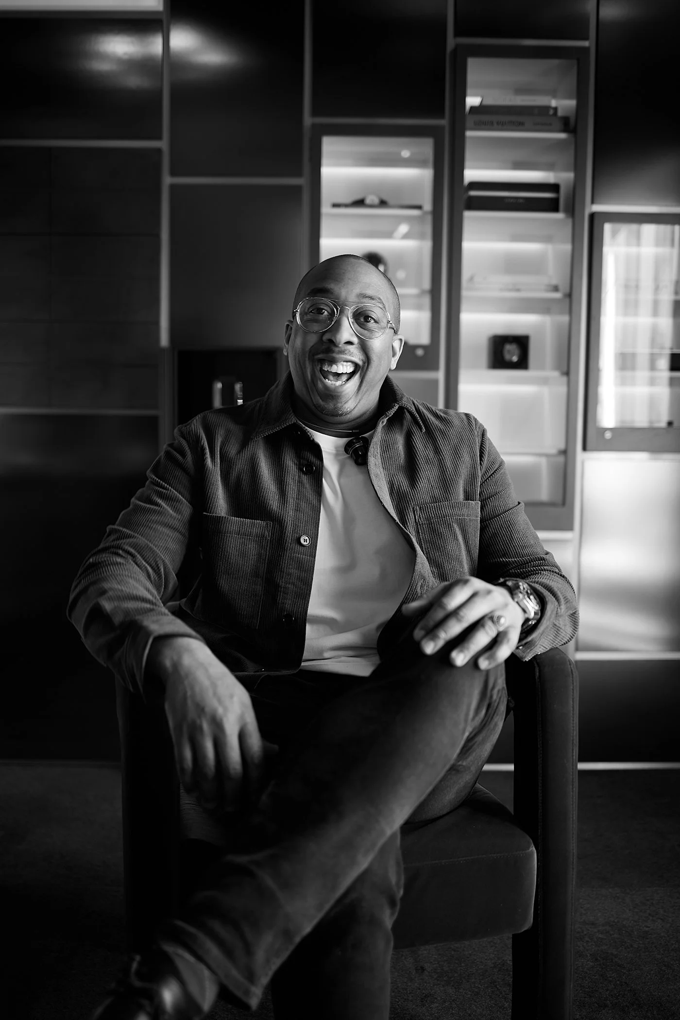 A smiling person with glasses sitting on a chair in a modern room with built-in shelves.