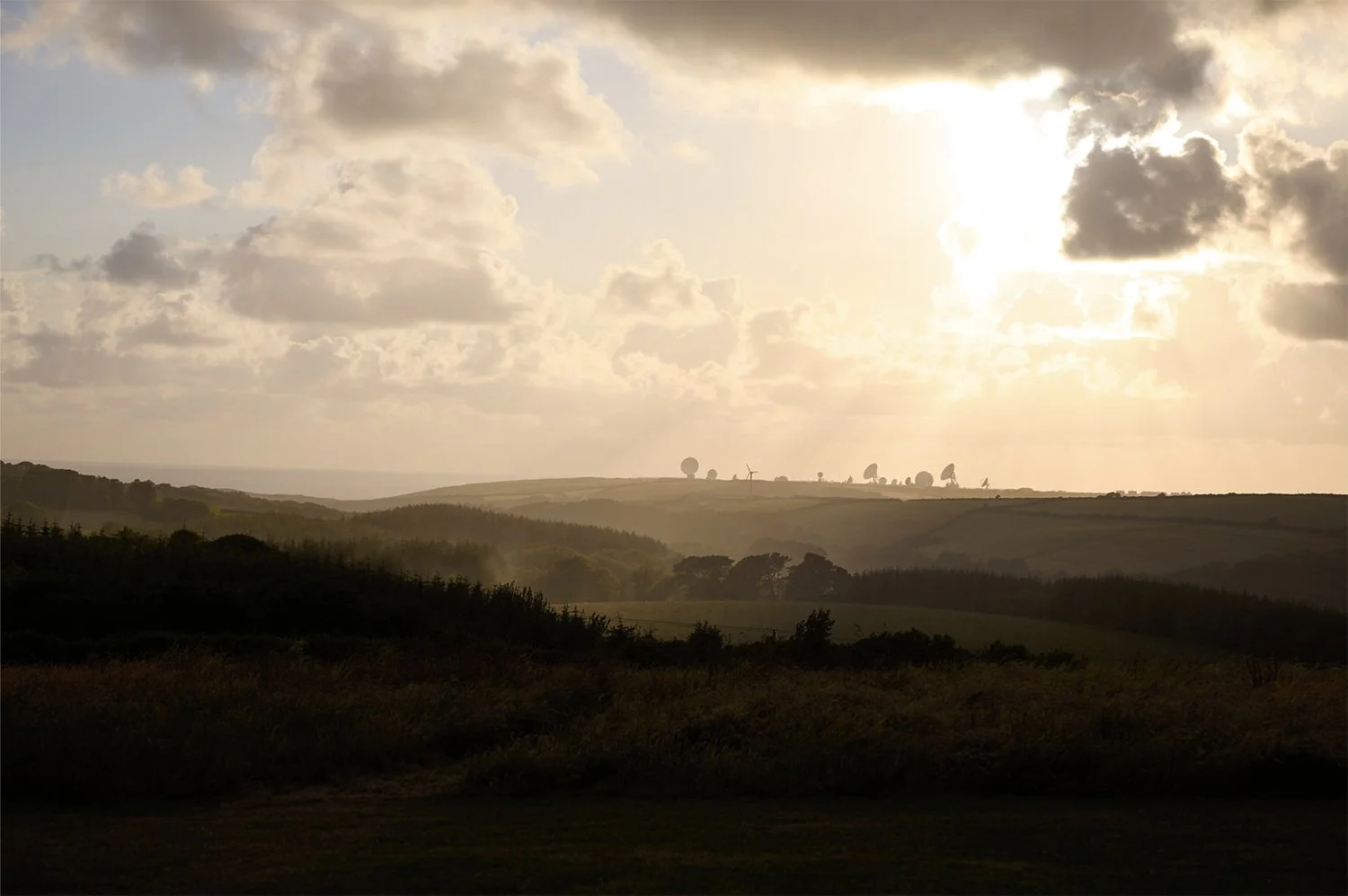 Sunset over a landscape with rolling hills, trees, and satellite dishes in the distance.