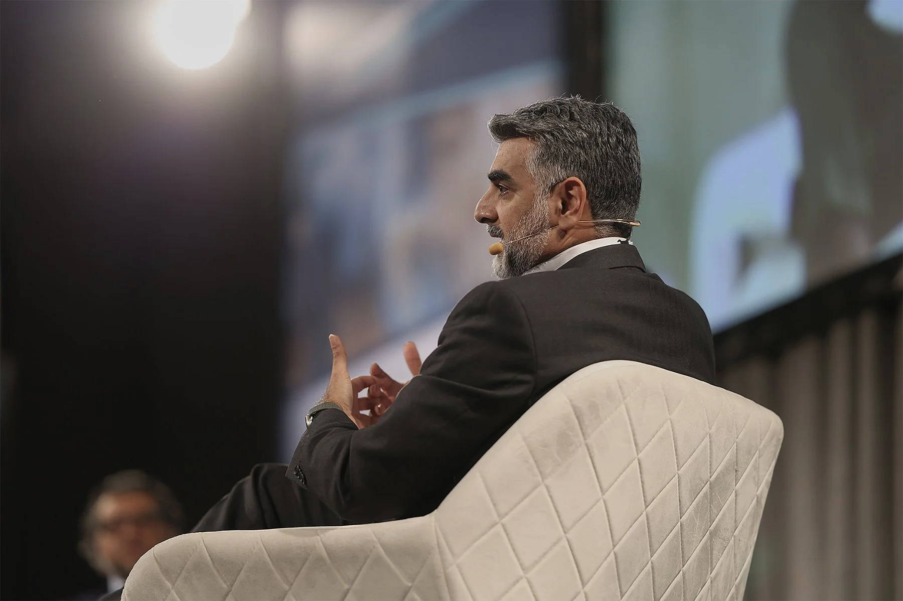 A man with gray hair and beard, wearing a dark suit, sits on a quilted white chair, speaking into a headset microphone during a conference or panel discussion.