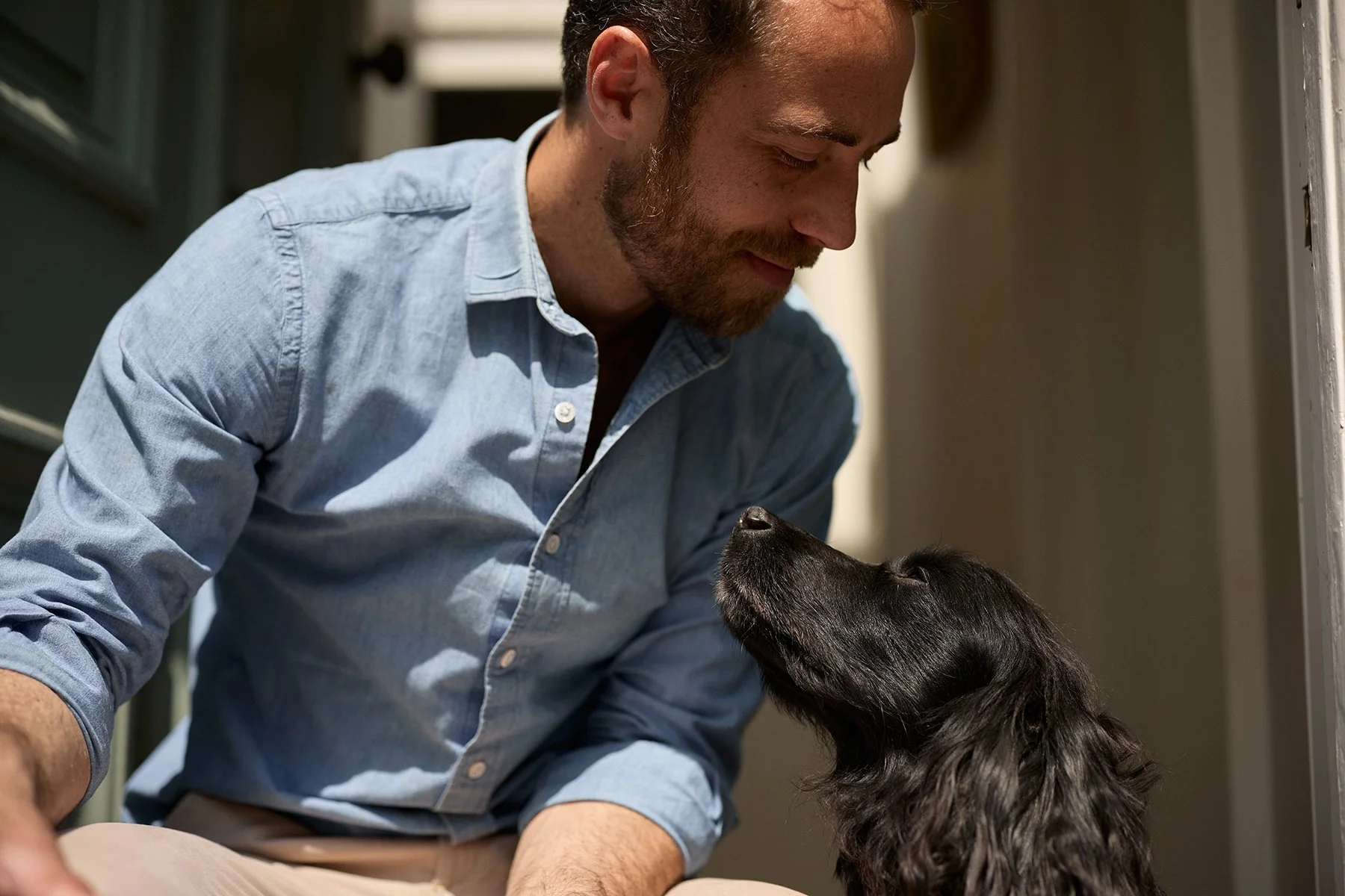 A man leaning down and smiling at a black dog with long, wavy fur, both looking at each other affectionately indoors.