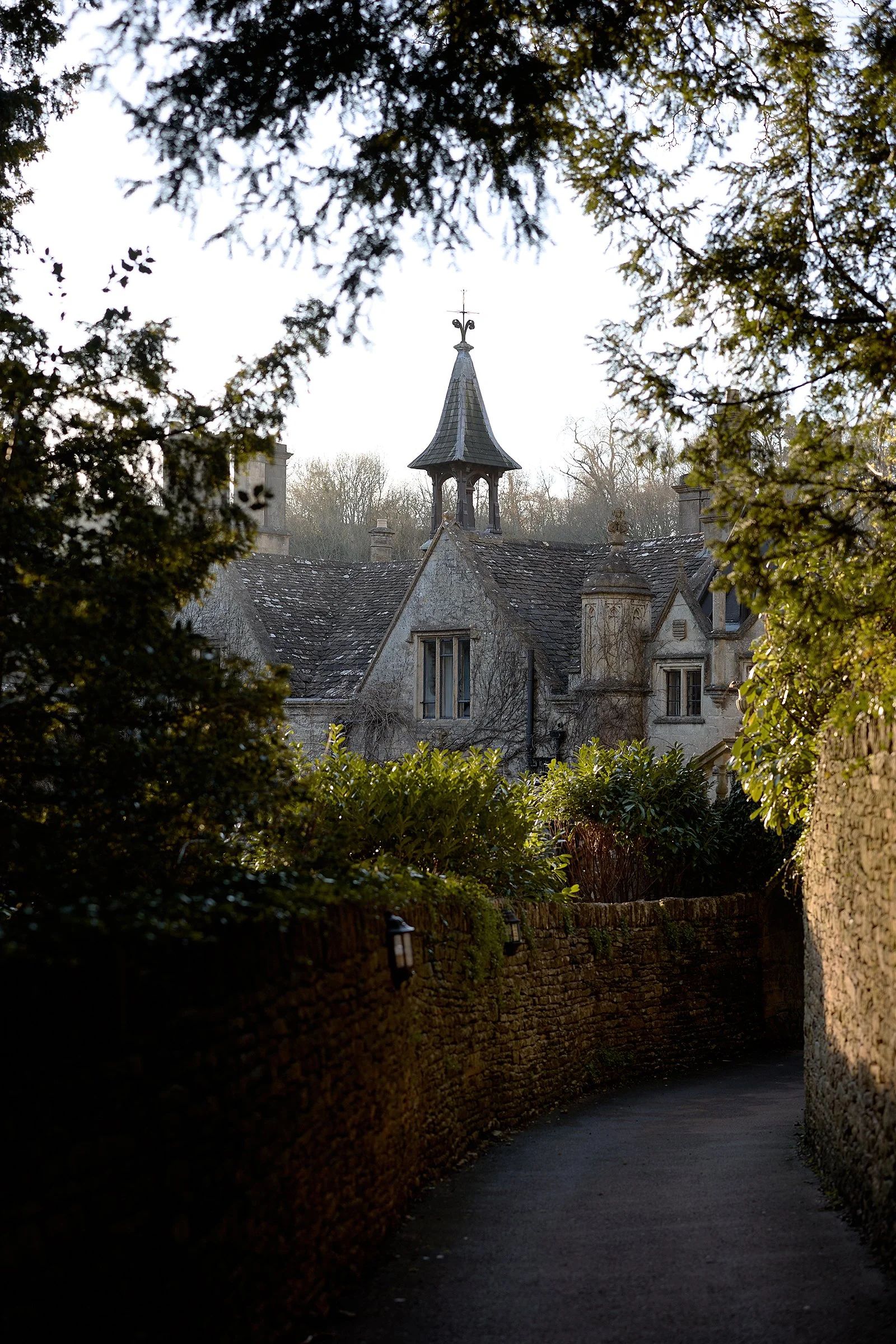 A narrow pathway bordered by stone walls leads to an old stone house with a steeply pitched roof and a small tower with a weather vane at the top, framed by trees.