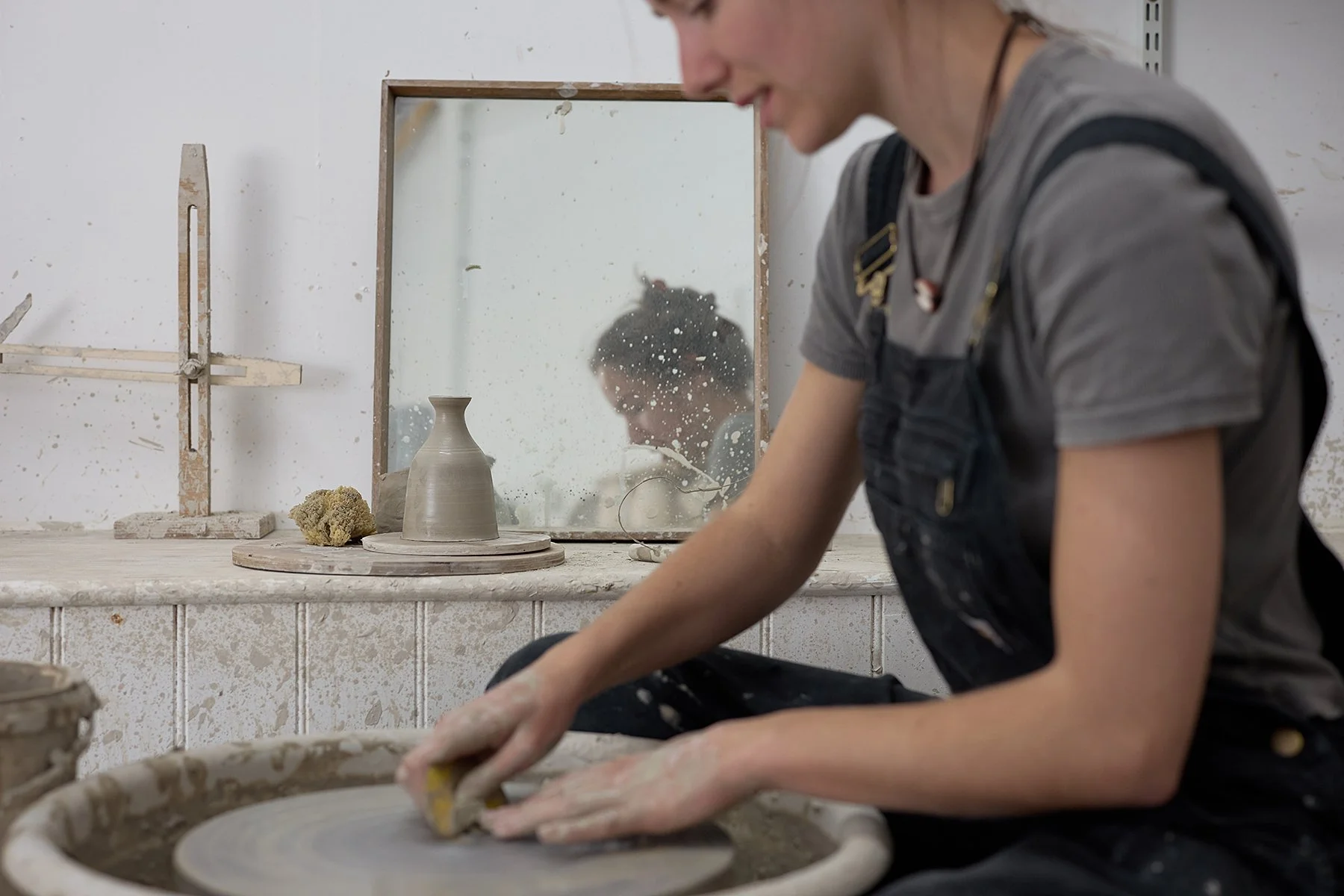 A woman shaping clay on a pottery wheel in a studio, with a mirror reflecting her focused face.